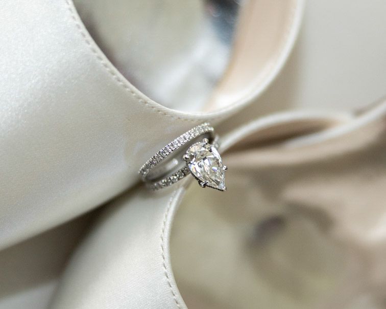 Pear-shaped diamond engagement ring with a pavé diamond band resting on cream satin bridal shoes.