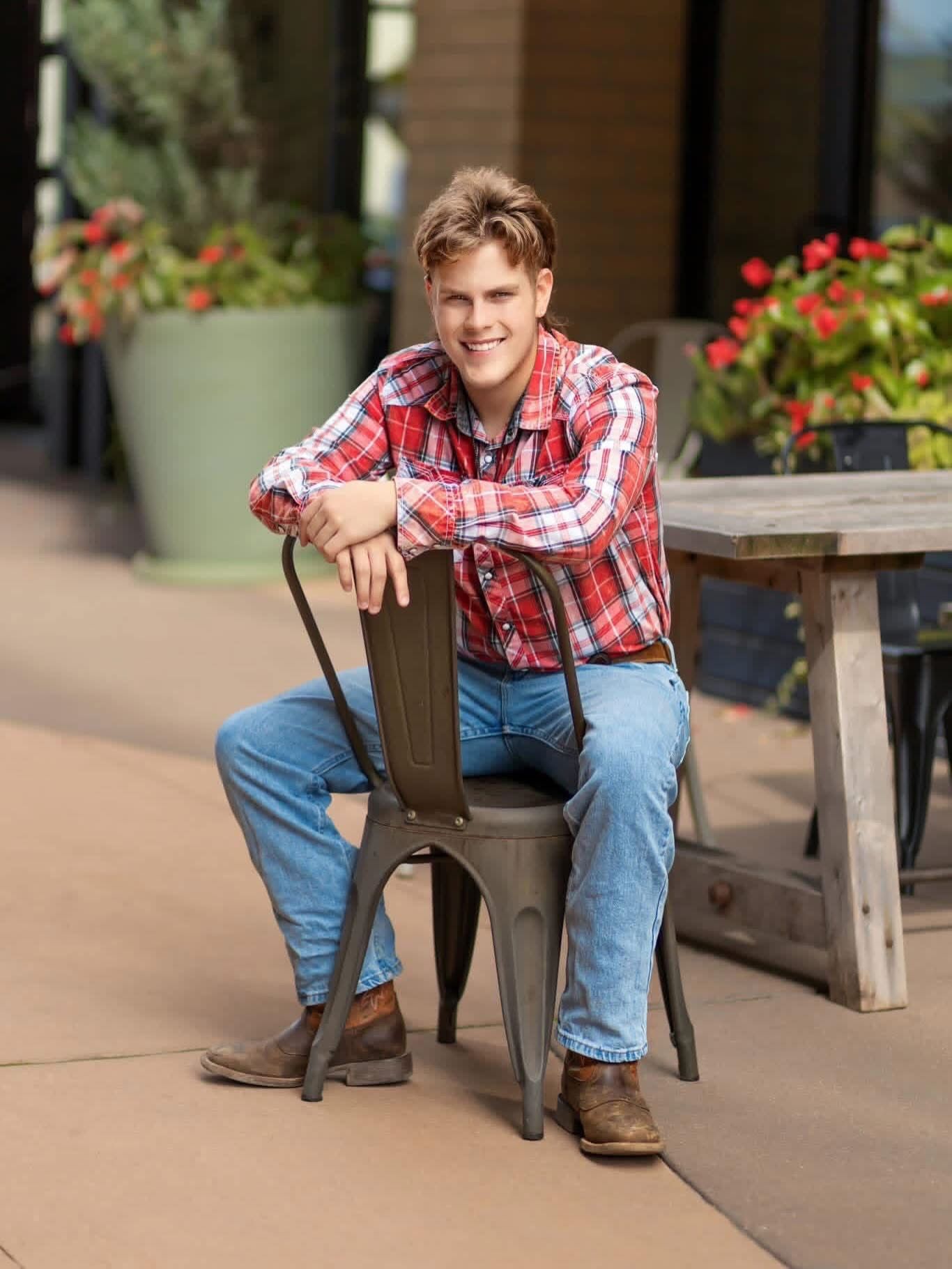 Smiling teenage boy in red plaid shirt and jeans sitting on a metal chair at an outdoor patio.