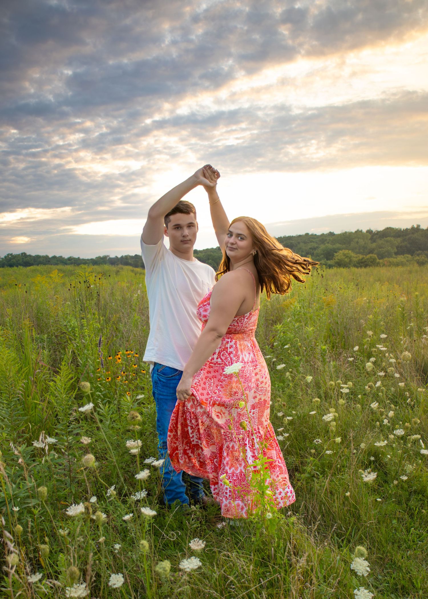 Young couple dancing in a wildflower meadow at sunset, woman in pink floral dress being twirled by man in white tee.