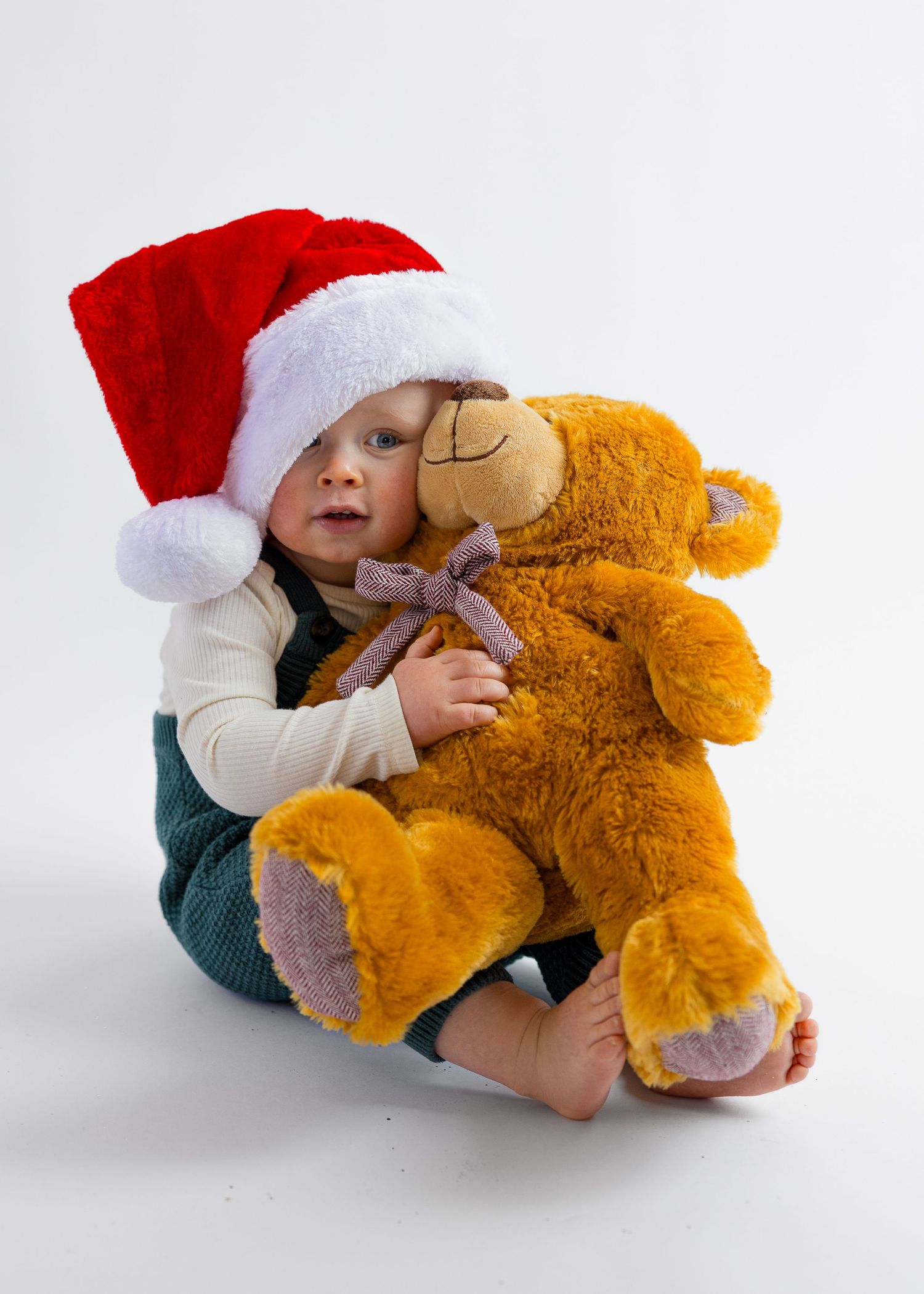 Baby in Santa hat cuddles a large brown teddy bear with a purple bow tie.