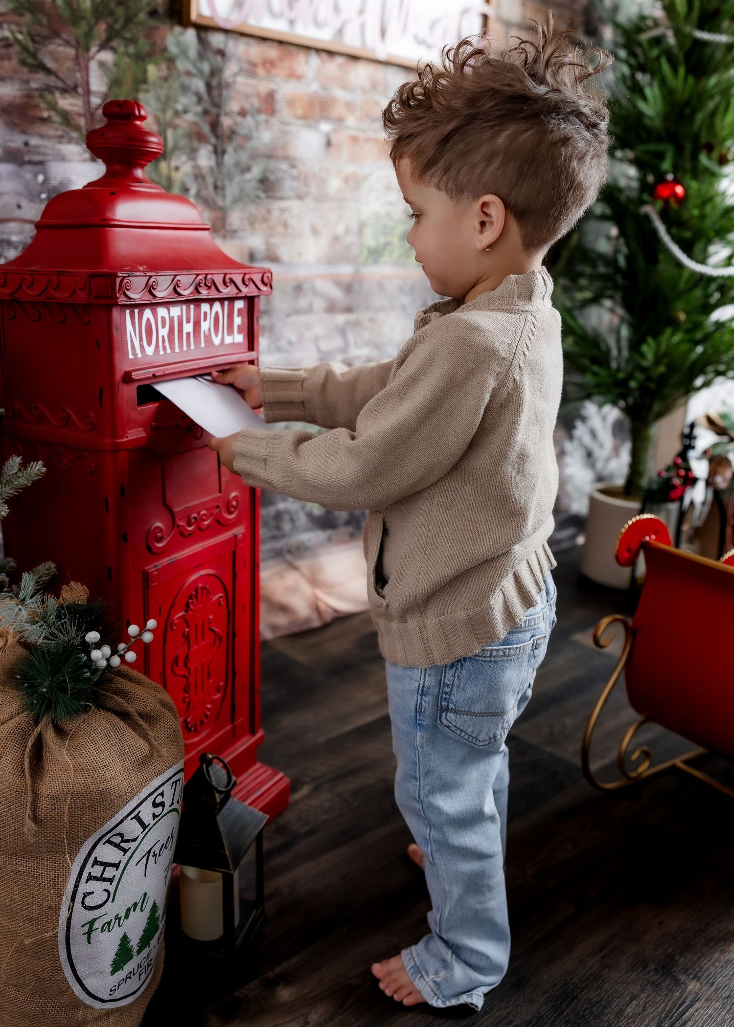 A toddler in a beige sweater and jeans mails a letter into a red North Pole mailbox in a Christmas scene.
