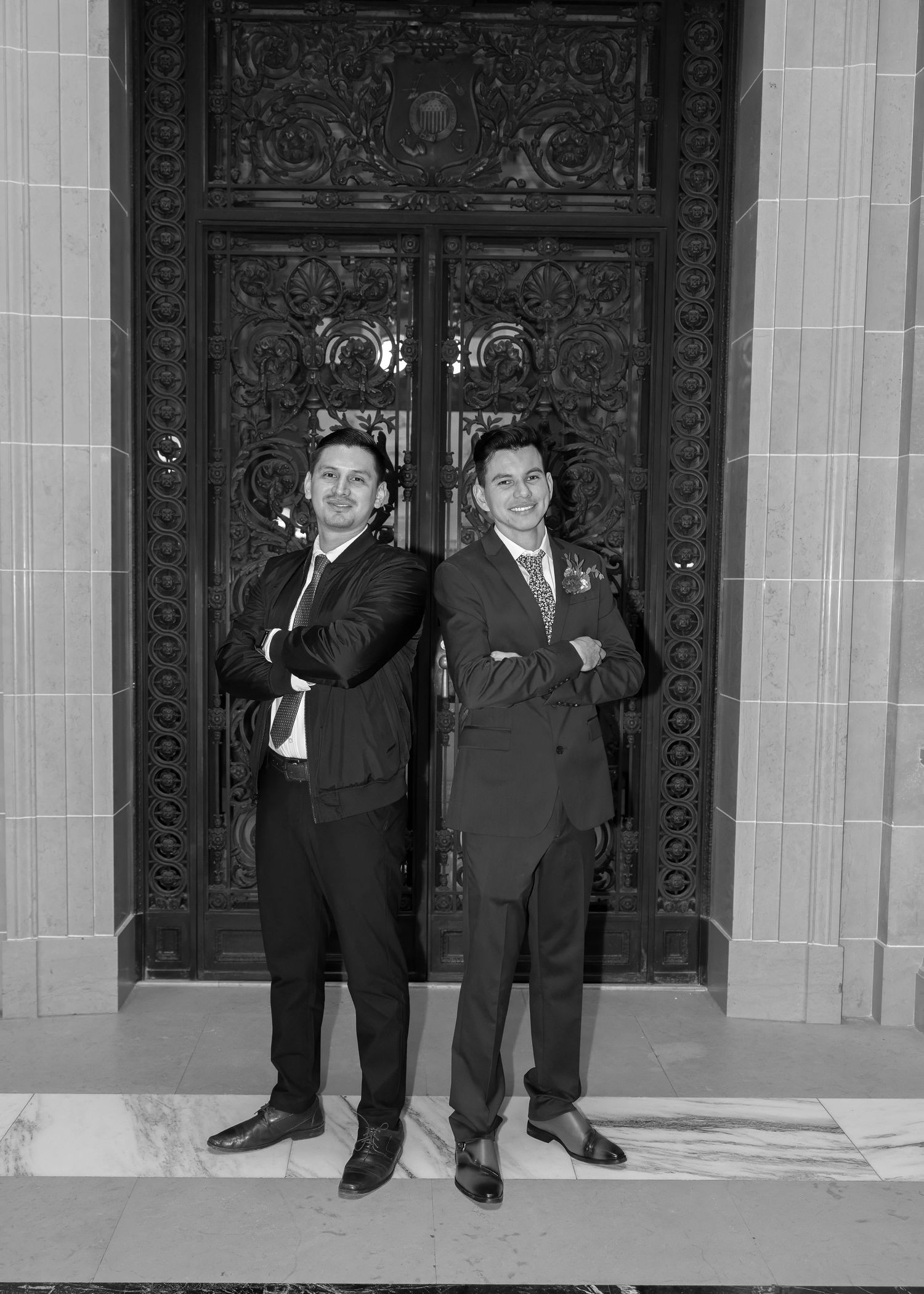 Two men in suits posing back-to-back before decorative doors in black and white wedding photo.