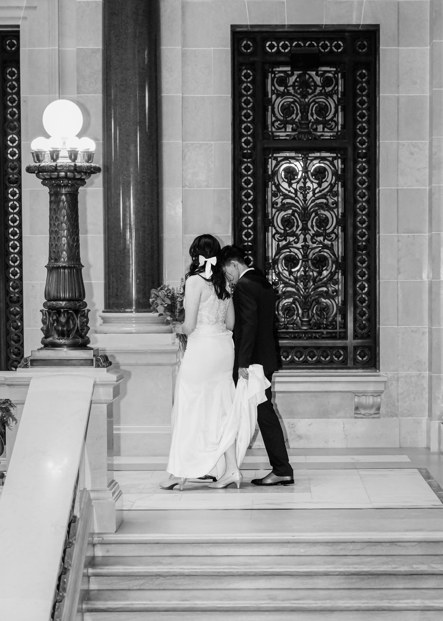 Black and white photo of wedding couple leaning together on marble steps by decorative door.