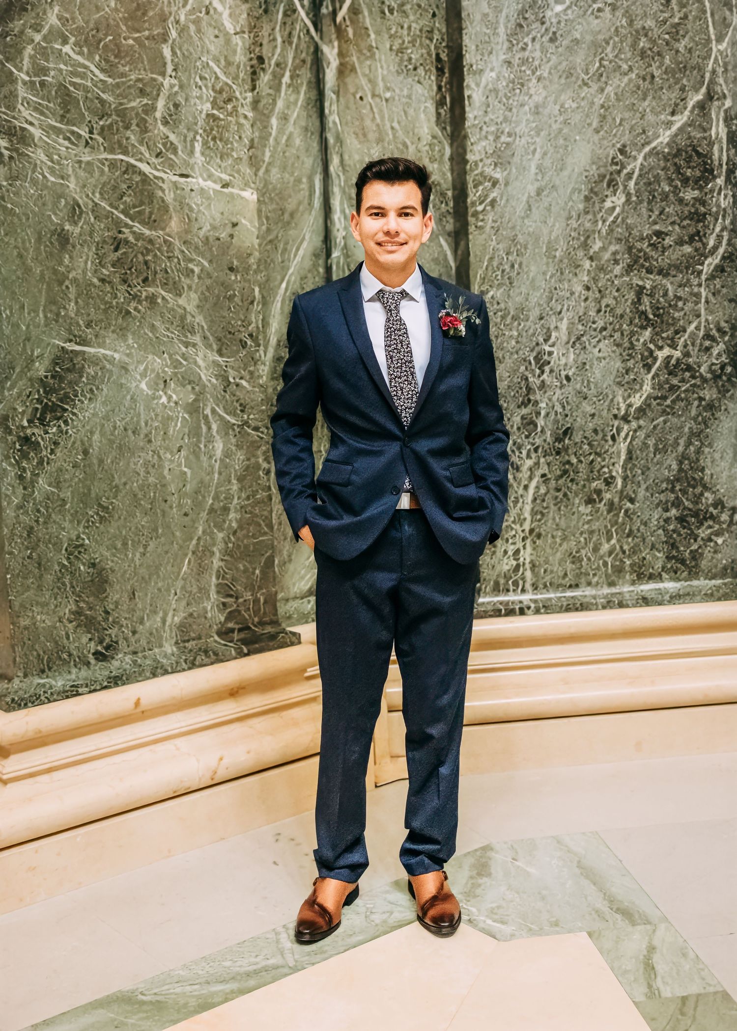 Groom in navy blue suit with floral boutonniere standing confidently in an elegant marble-walled venue.