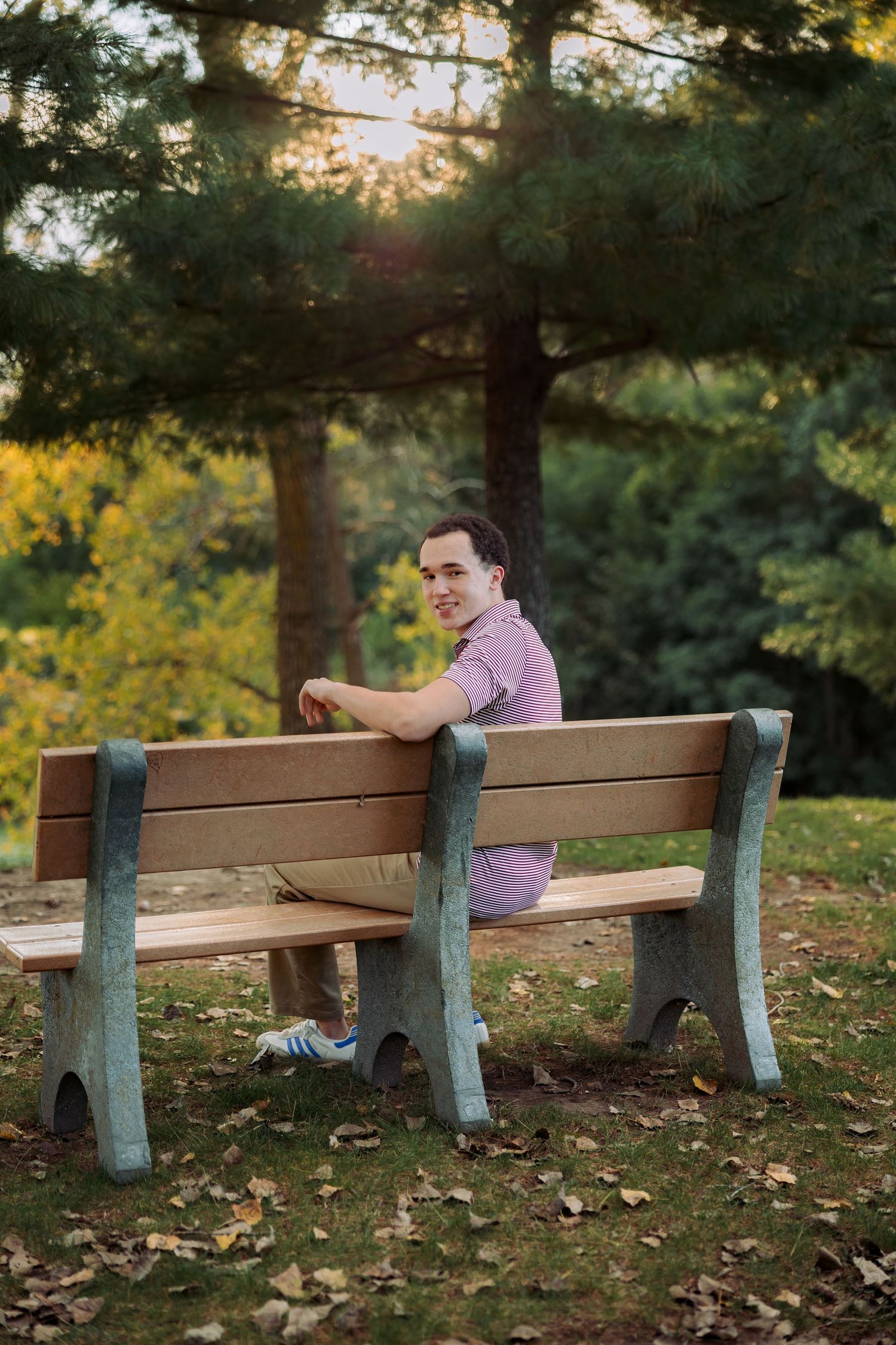 Young man in pink polo shirt sitting on a wooden park bench surrounded by lush green trees in golden hour light.