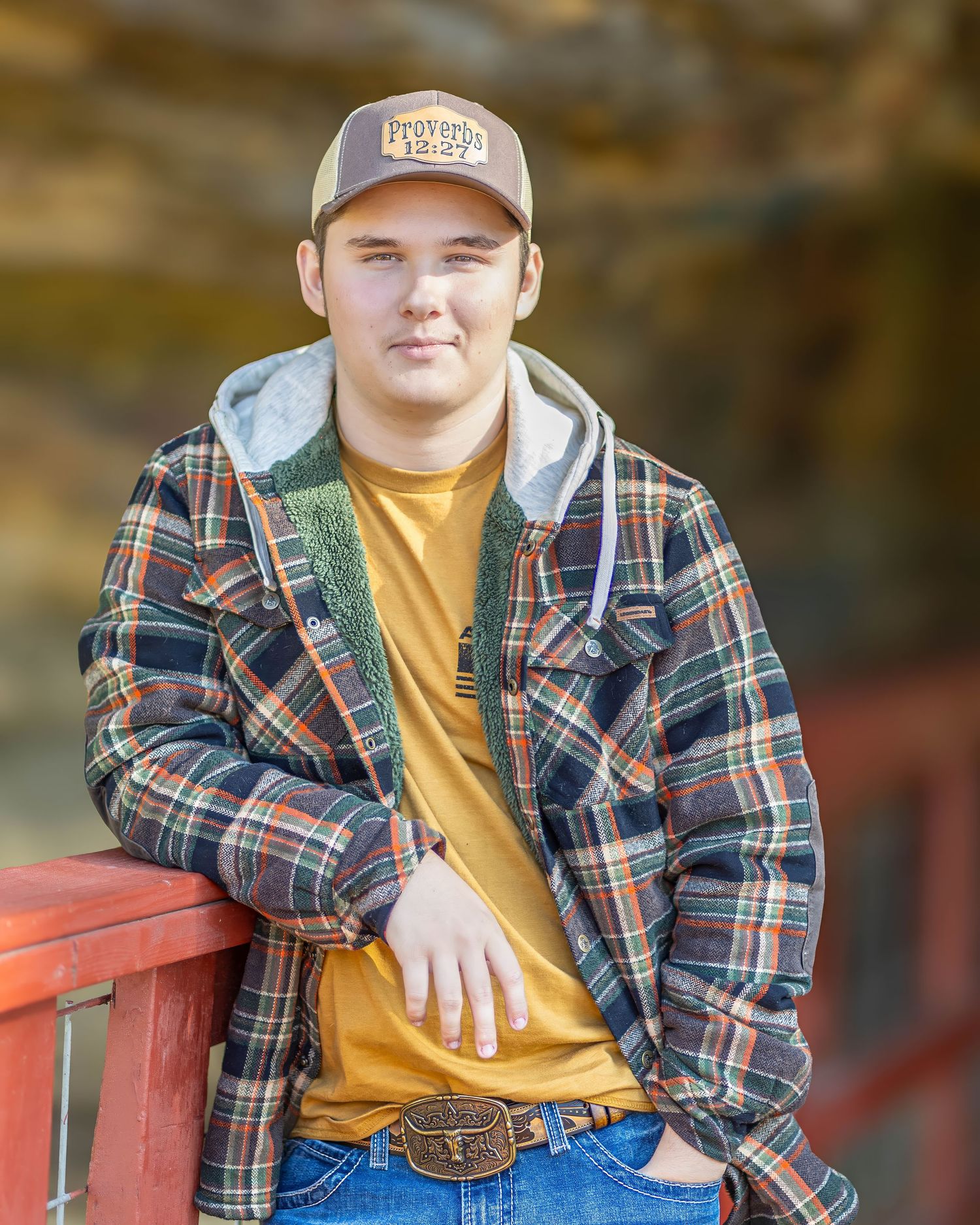 Teen boy in plaid jacket and trucker hat leans on red fence for fall senior photos.