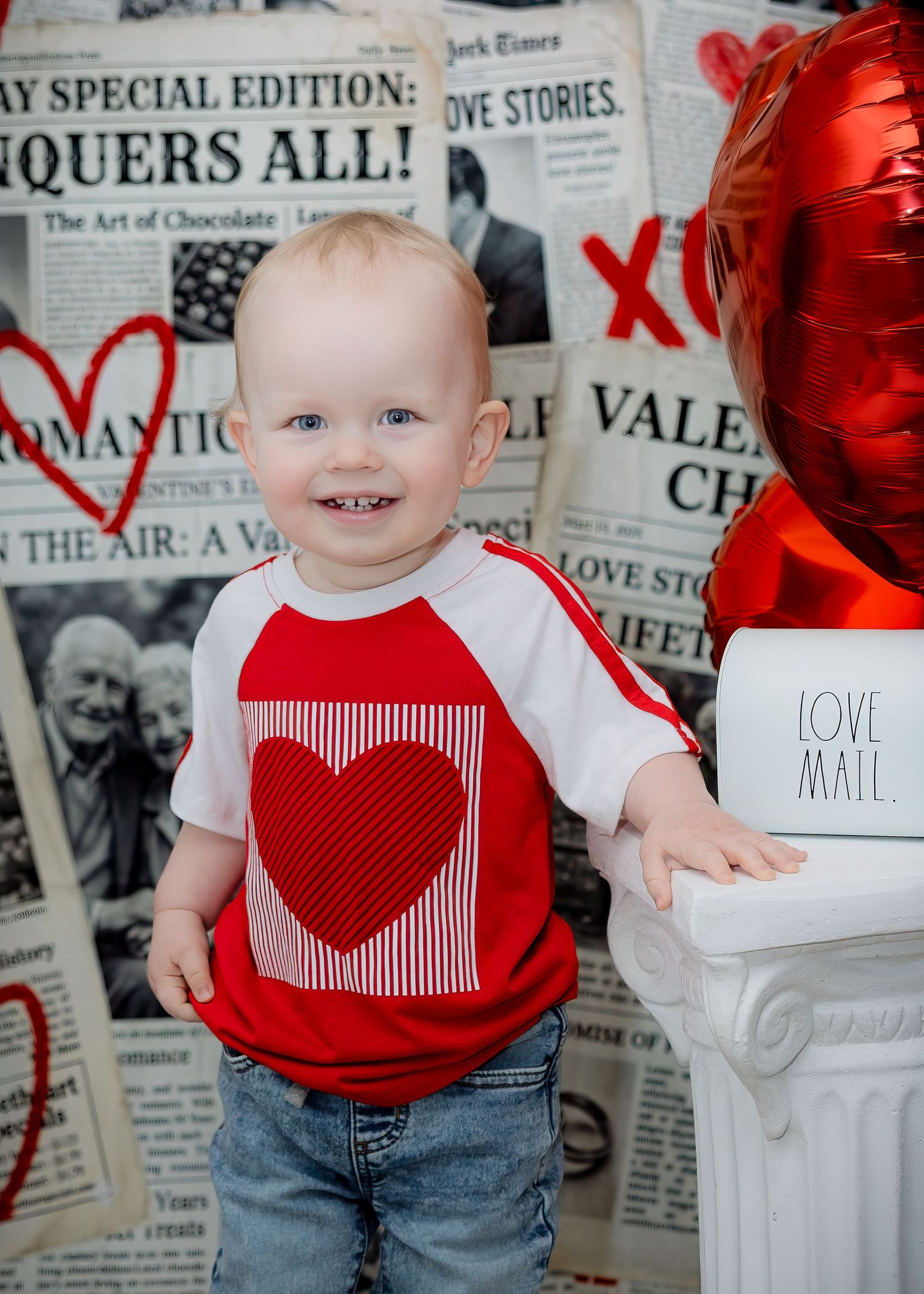 Smiling baby in red heart shirt poses with love mail box and balloon.