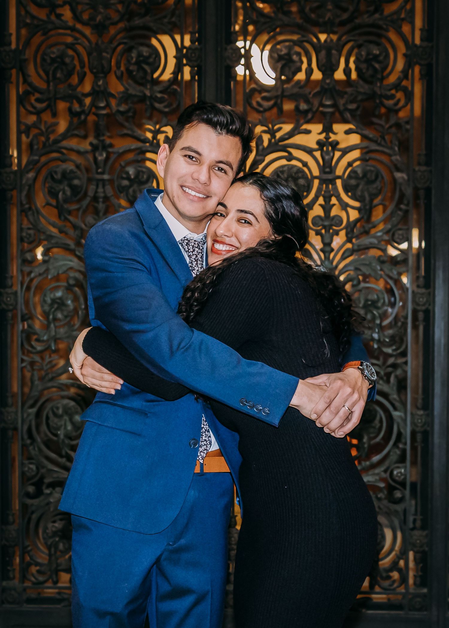 Groom in blue suit shares a joyful embrace with a smiling female guest before ornate iron doors.