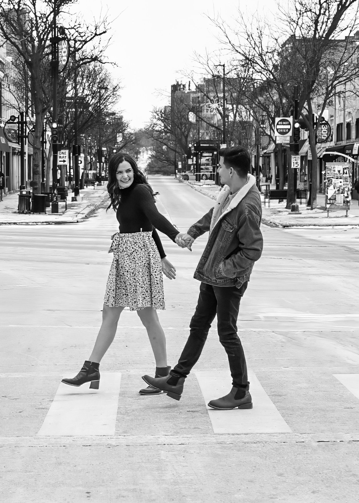 Black and white photo of couple holding hands crossing a zebra crosswalk on a wide city street.