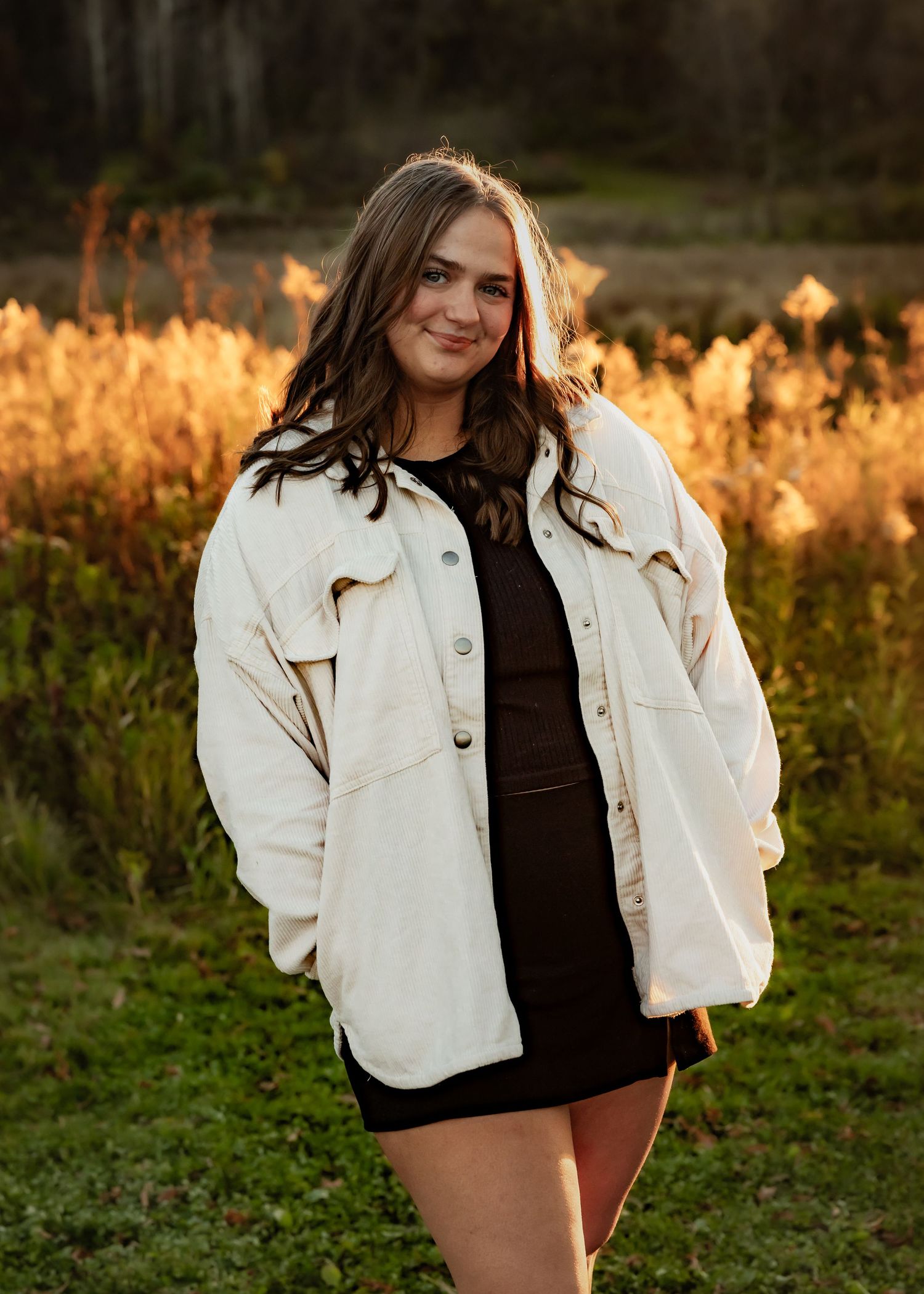 Young woman in cream jacket and black dress poses in golden field at sunset.