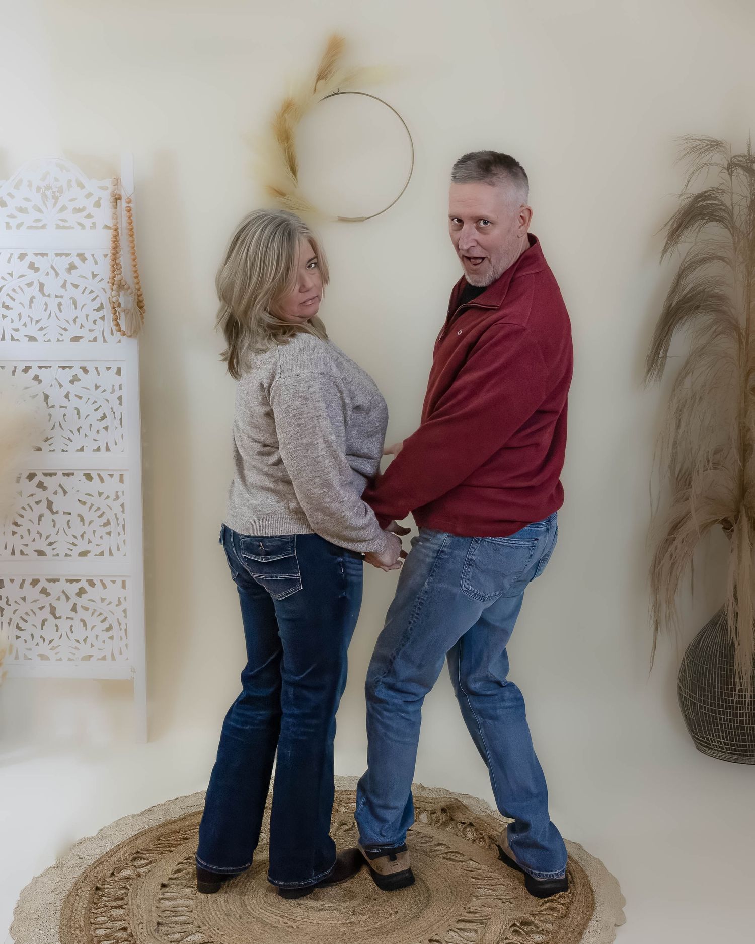 A laughing middle-aged couple holding hands and dancing playfully in a bright boho-styled studio.