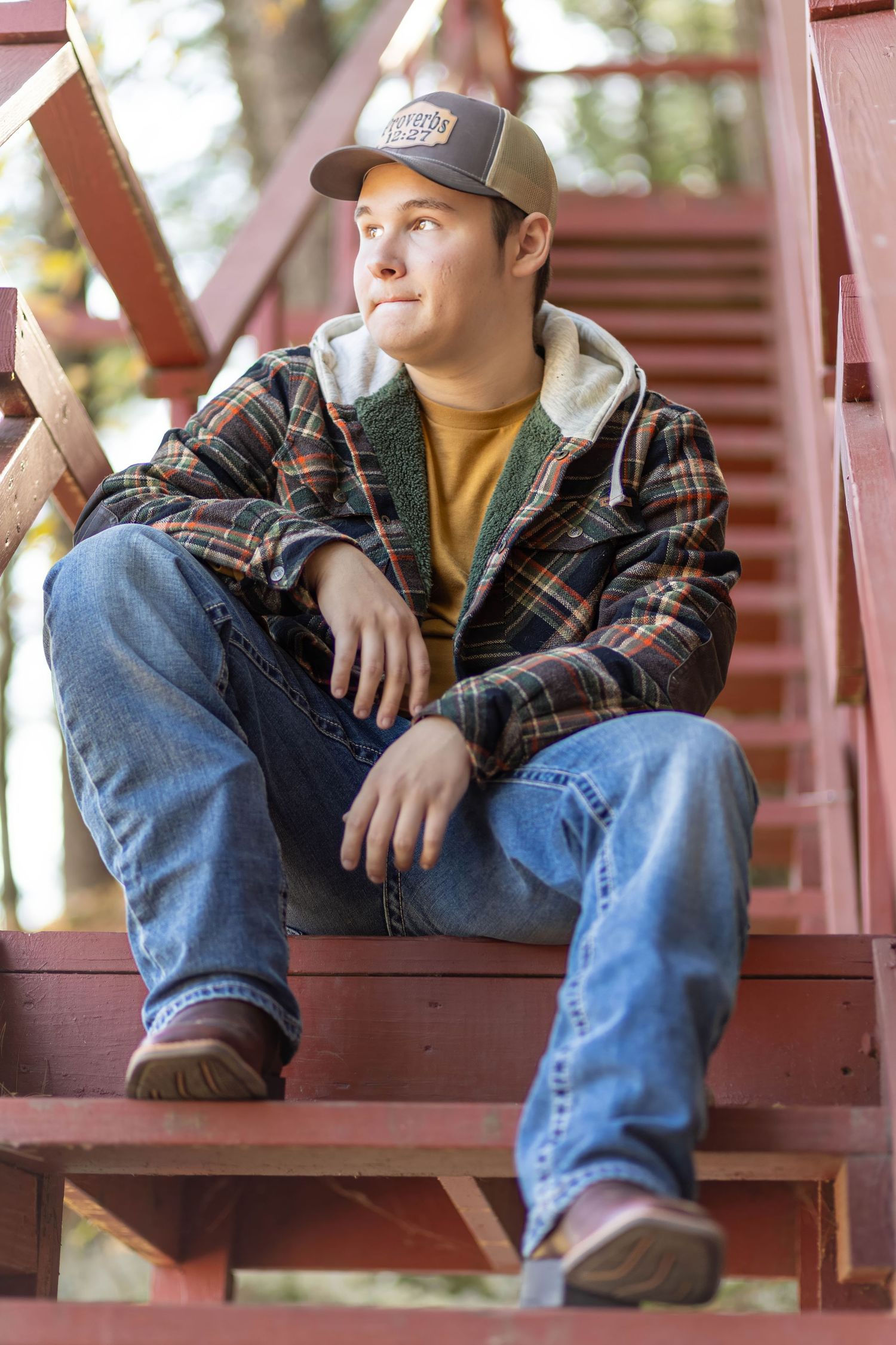 Young man in plaid jacket and cap sits on red wooden stairs for outdoor senior portrait.
