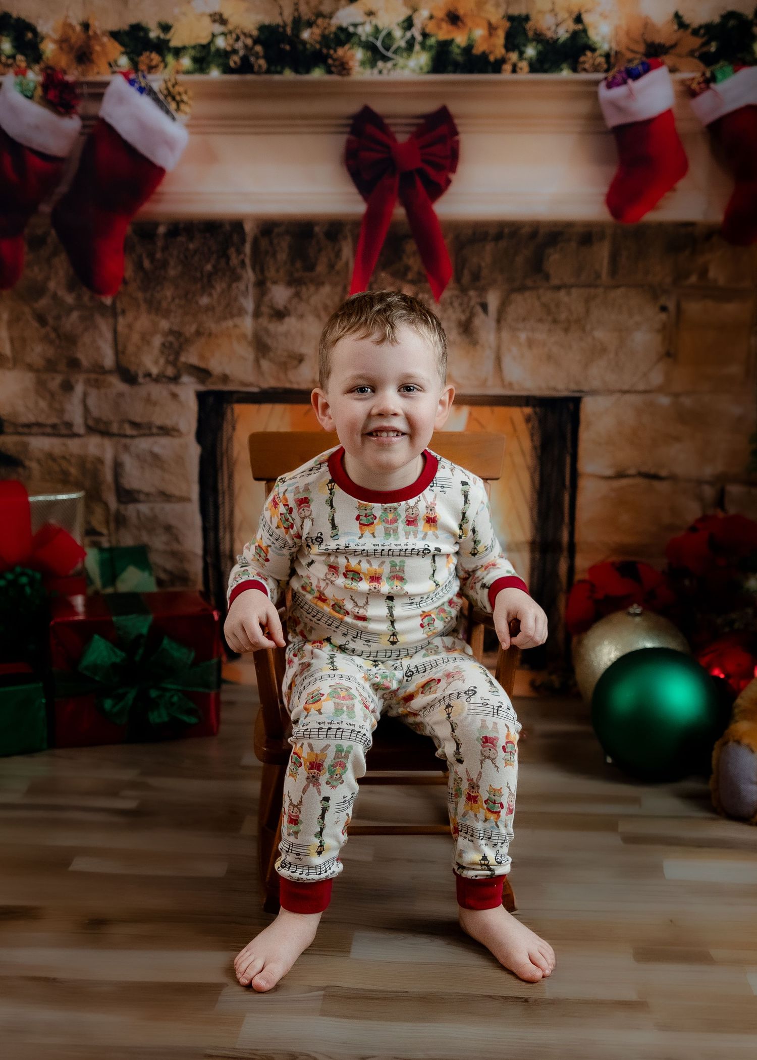 Smiling toddler in Christmas pajamas sitting on chair by festive fireplace with stockings and gifts.