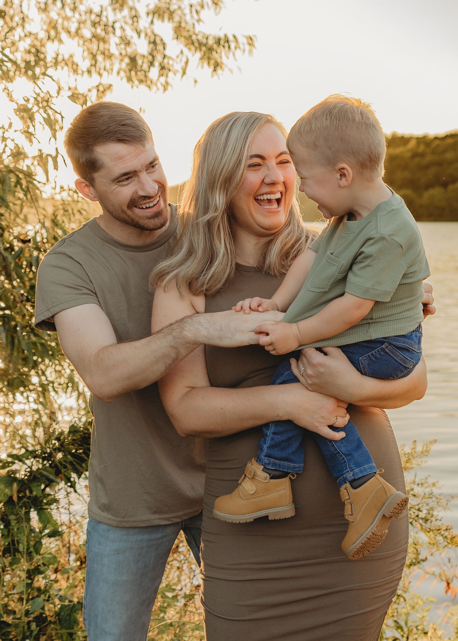 A laughing family of three enjoys a golden hour outdoor moment as mom holds their toddler son.