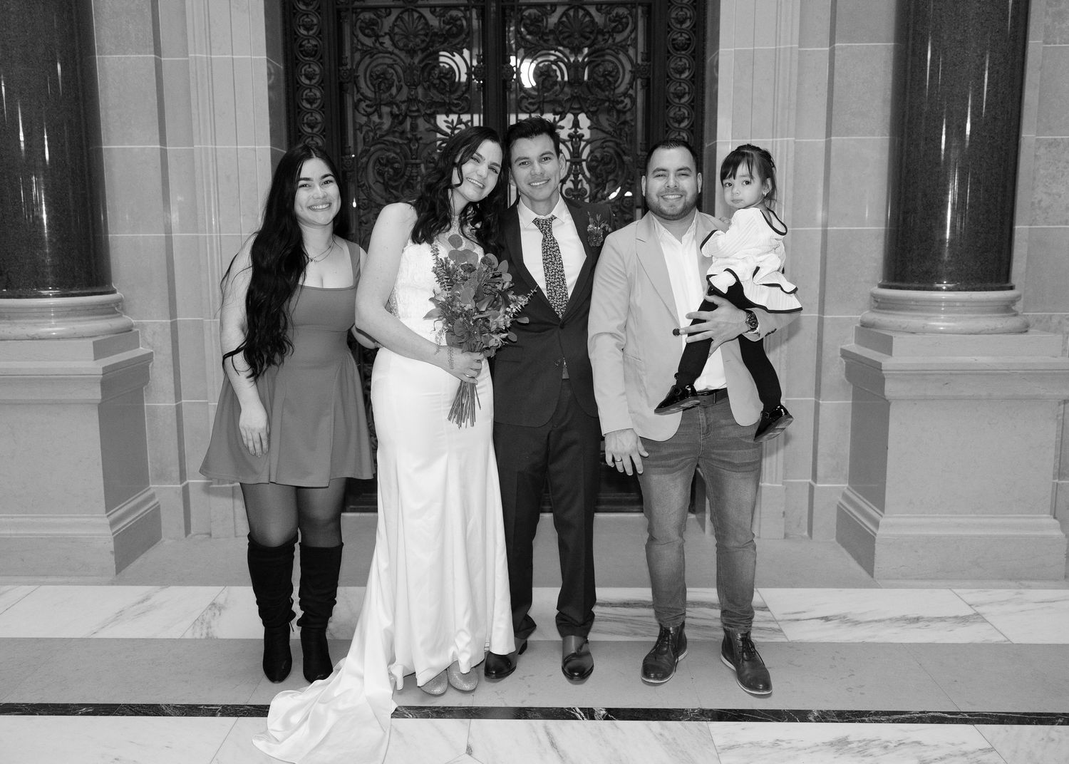 A wedding party of five, including a bride and groom, poses before ornate doors in black and white.