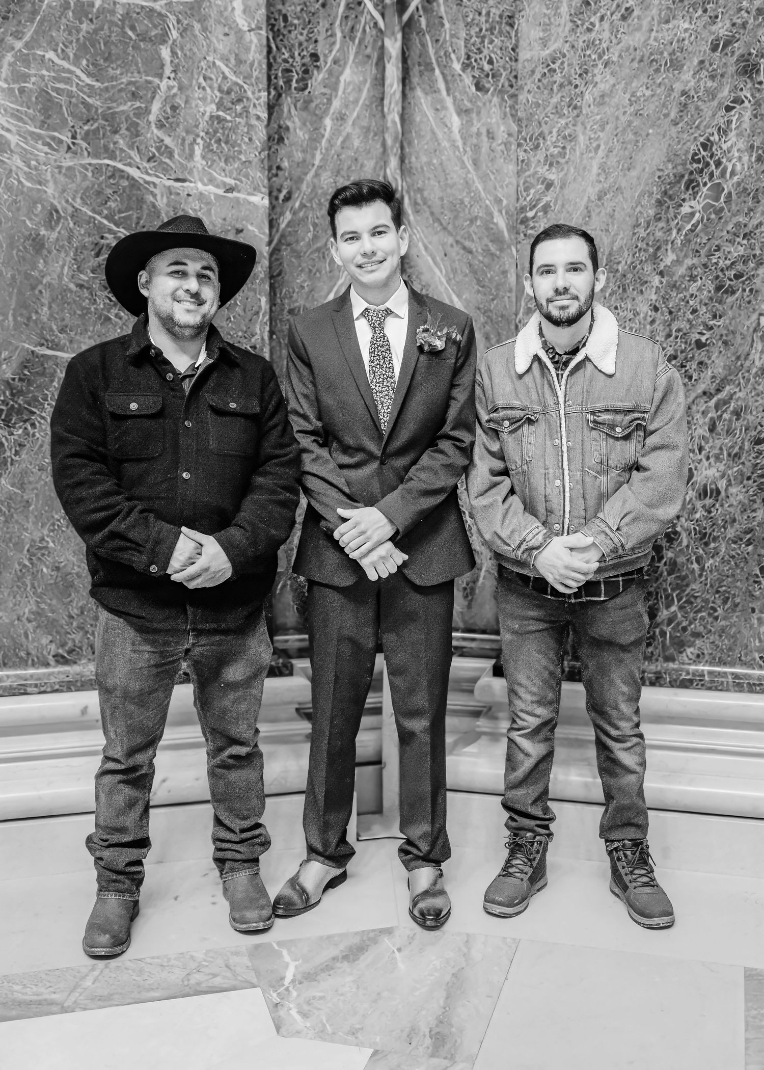 Groom in suit posing with two casually dressed men flanking him in a grand marble building, black and white.