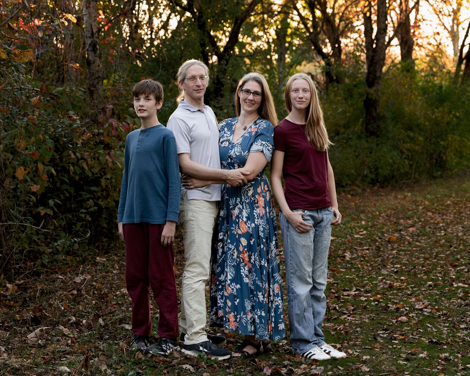 Family of four in casual clothes standing close together on a leaf-covered path in a sunlit autumn park with trees behind them.