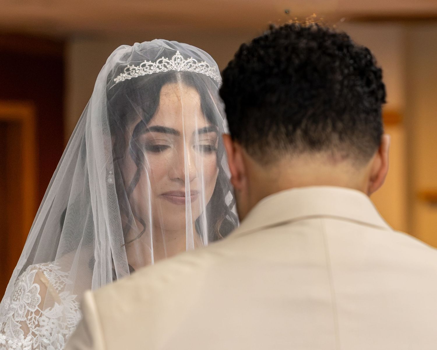 Bride in a lace veil and sparkling tiara gazes at the groom, who faces away in a cream suit, during a wedding ceremony.