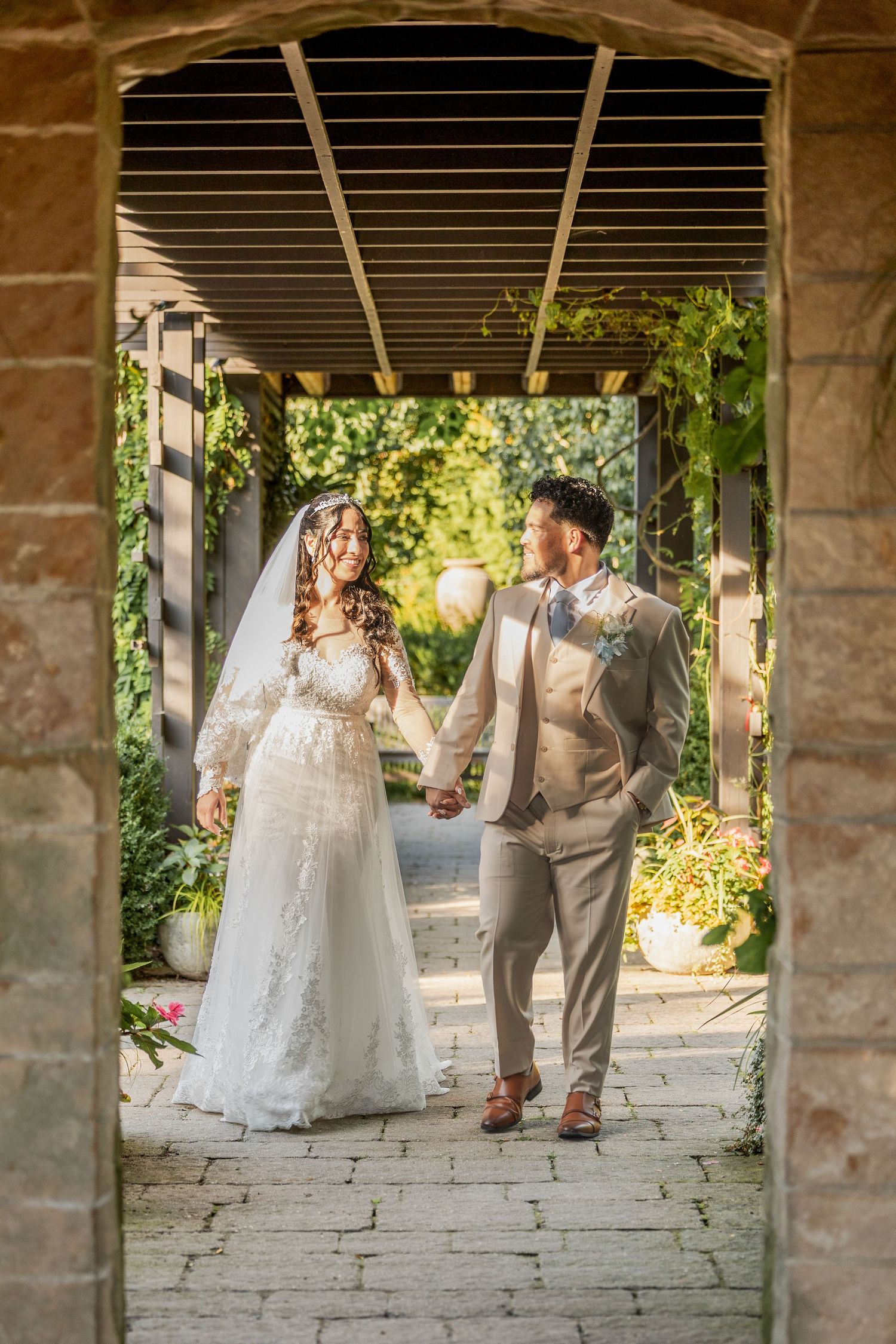 Smiling bride in a lace wedding gown and groom in a tan suit hold hands while walking through a sunlit garden pergola archway.