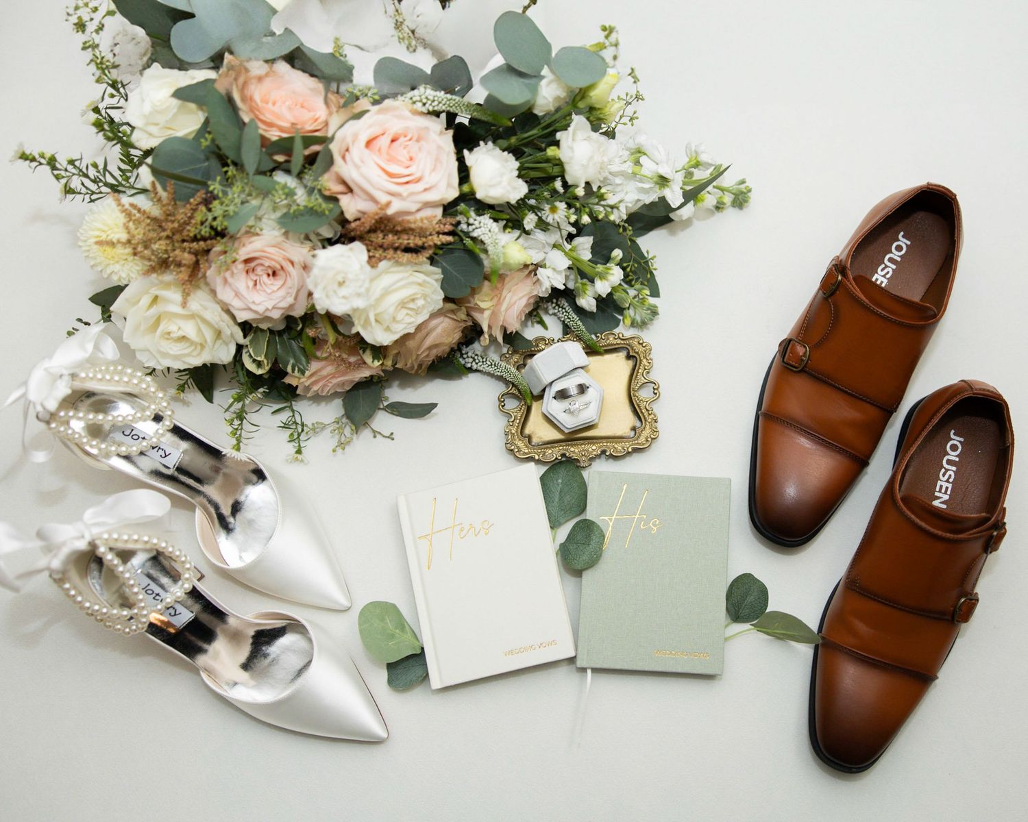 Wedding flat lay with a blush rose bouquet, white bridal heels, brown leather groom shoes, and two vow books on a white surface.
