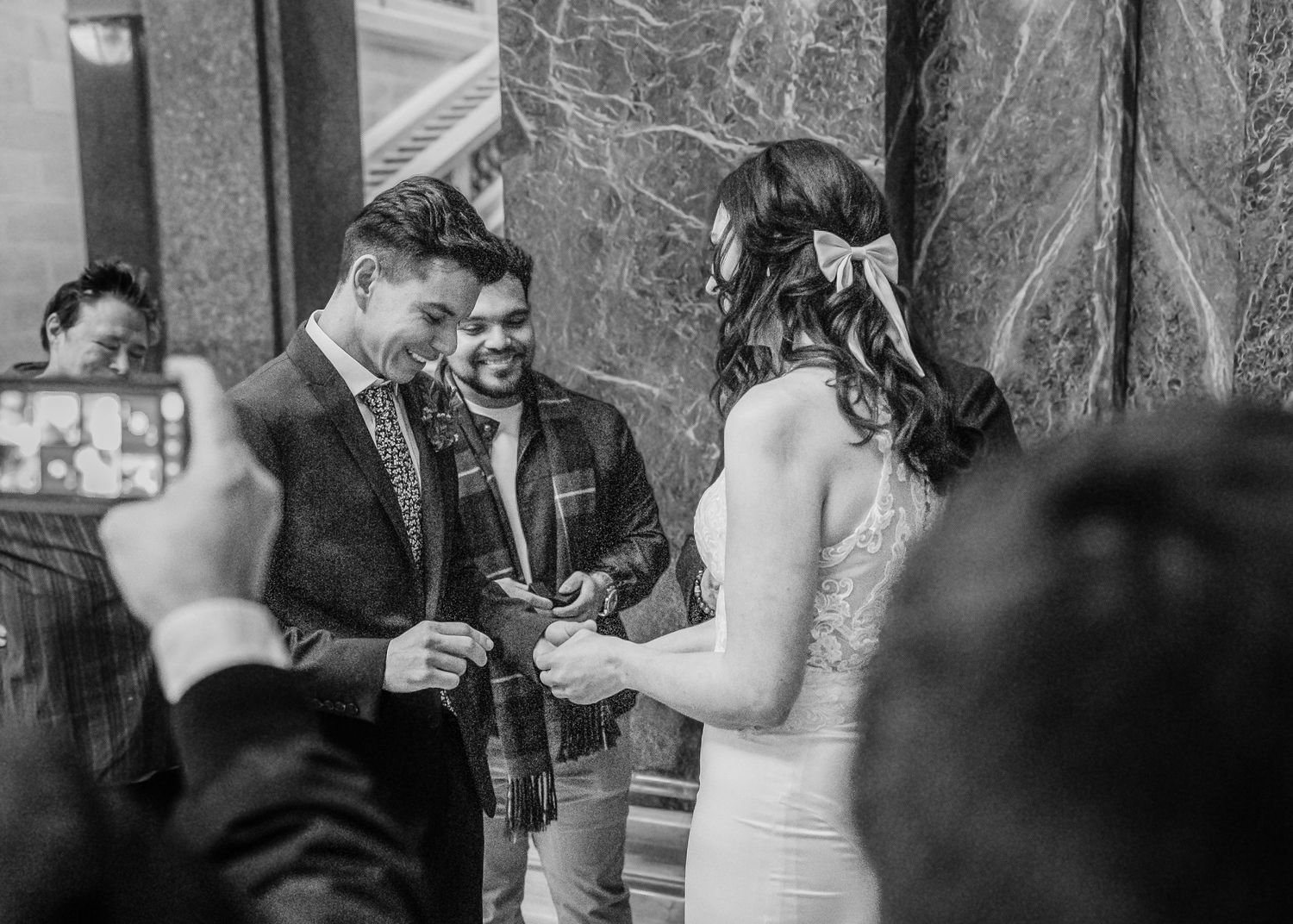 Groom placing wedding ring on brides finger while guests watch and photograph the moment.