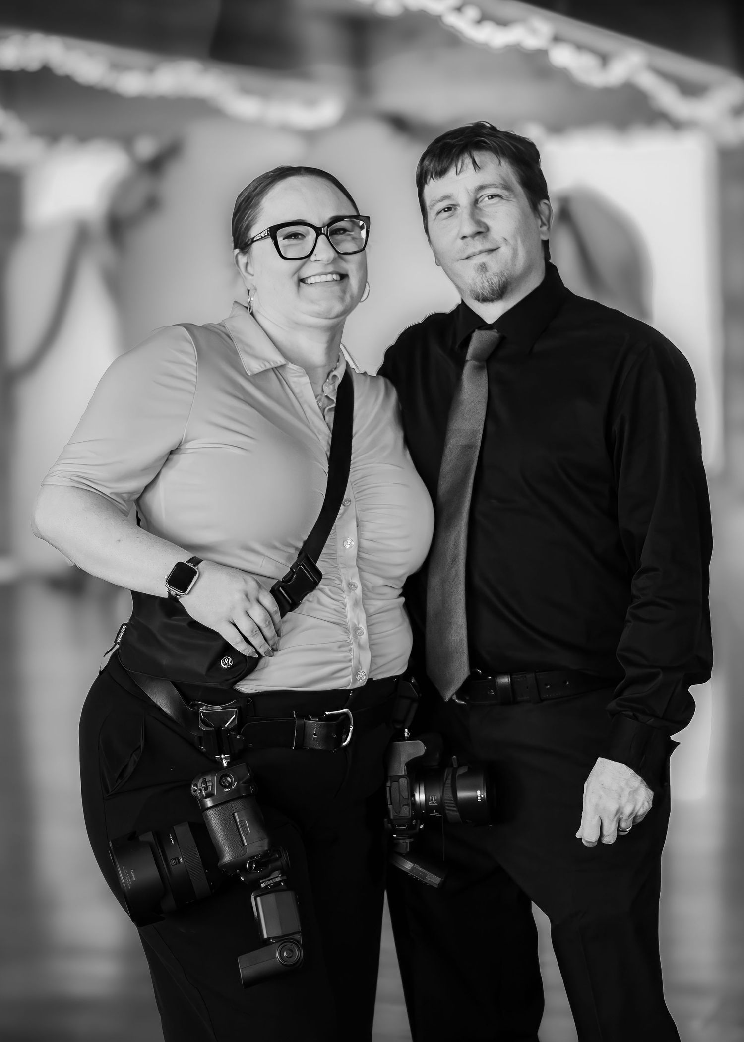 Two professional photographers smile together holding cameras in a black and white portrait.