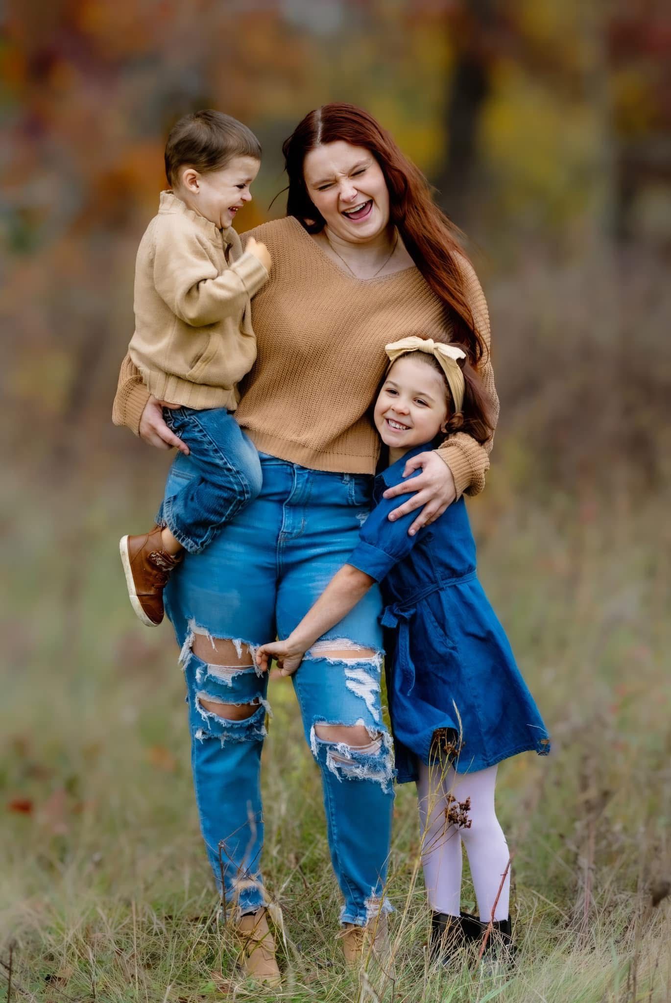 Laughing redhead mother in tan sweater and ripped jeans hugs two young children in autumn field.