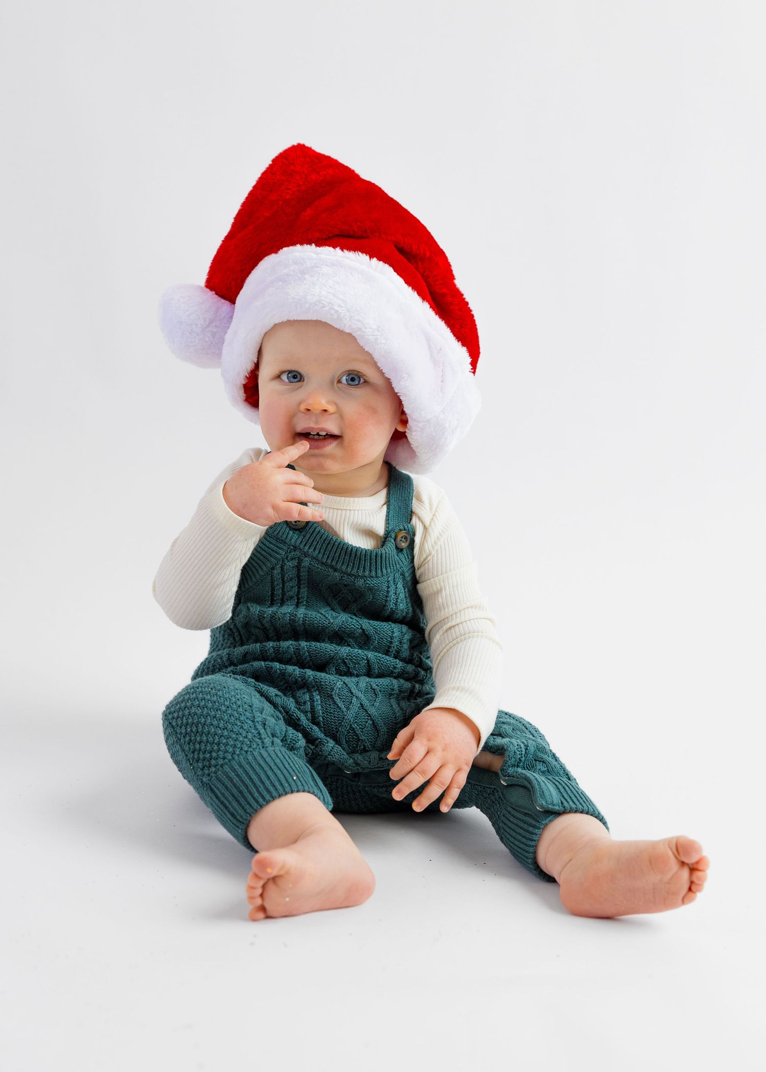 Cute baby wearing Santa hat sits barefoot in green knit overalls chewing on a finger.