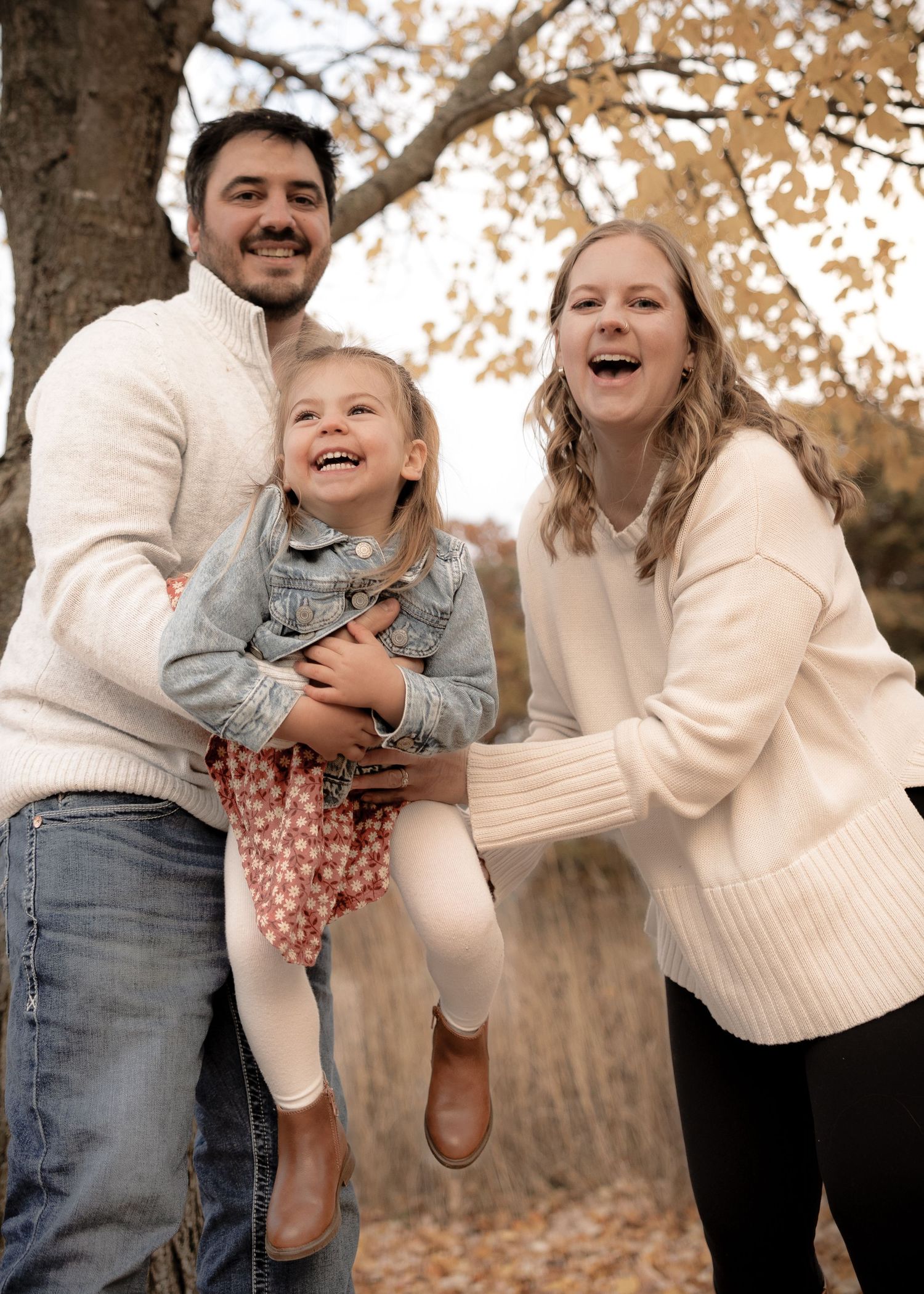 Happy family of three laughs together outdoors beneath golden autumn foliage.