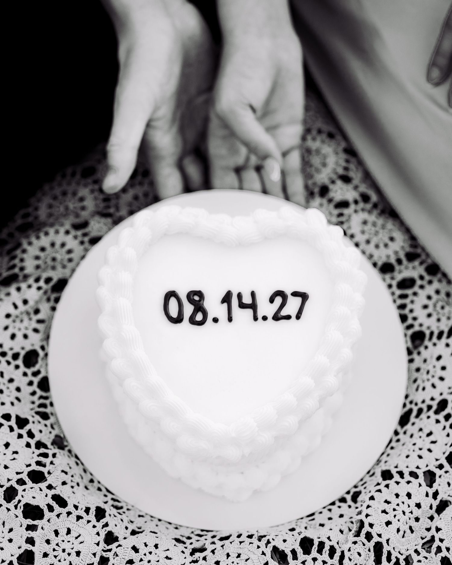 Black and white photo of hands holding over a heart-shaped white cake with date 08.14.27 on lace tablecloth.