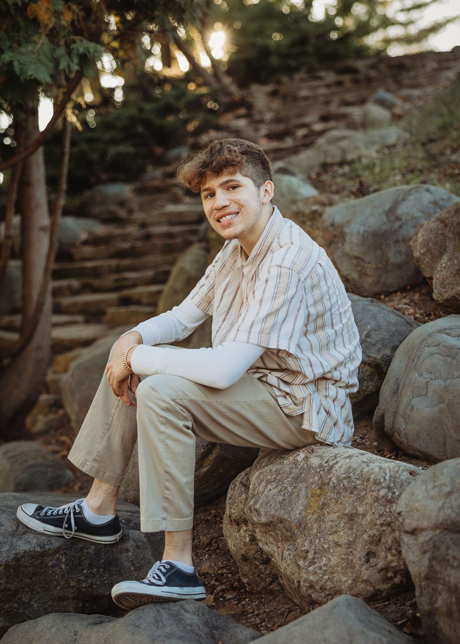 Young man smiling, sitting on rocks outdoors, wearing striped shirt, khaki pants, and Converse sneakers, with stone steps behind.
