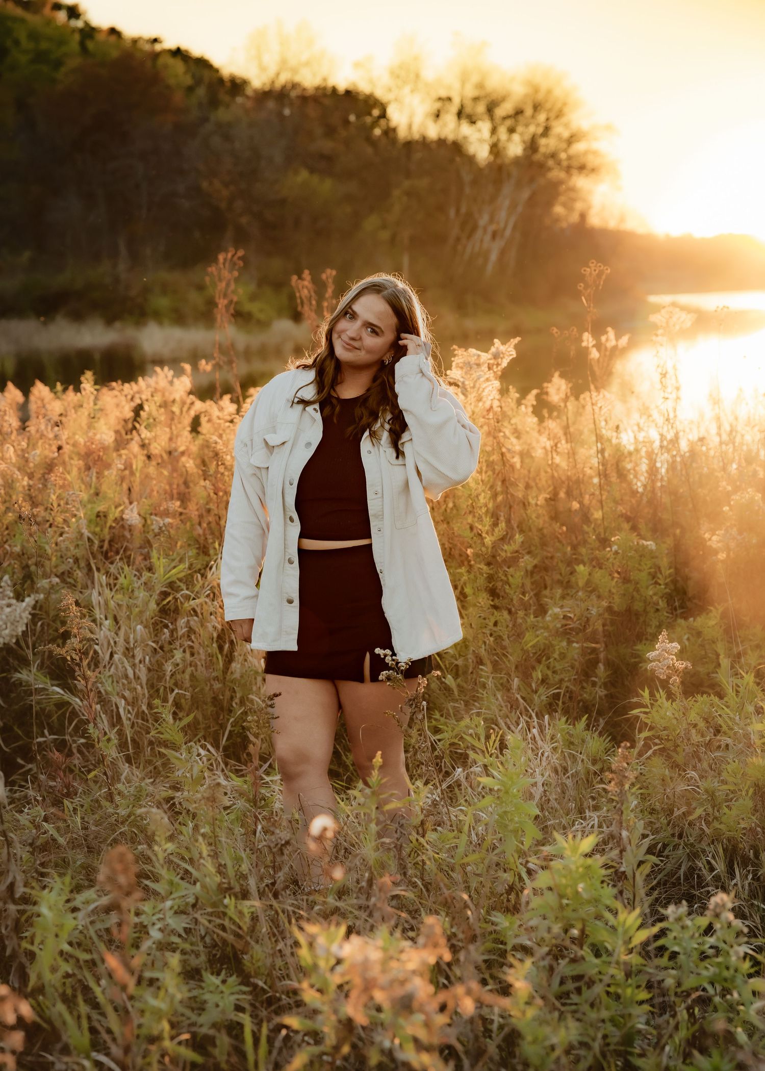 Young woman in white jacket and dark skirt standing in golden field at sunset near a lake.