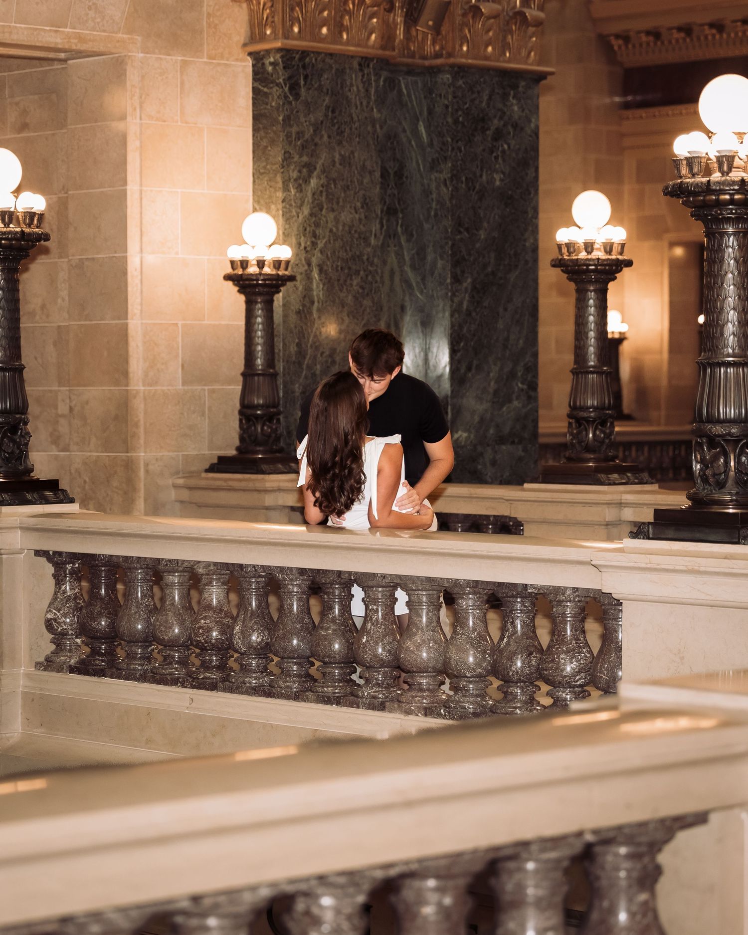 Couple sharing a romantic kiss inside an elegant marble building with ornate lamp posts and grand balustrade.