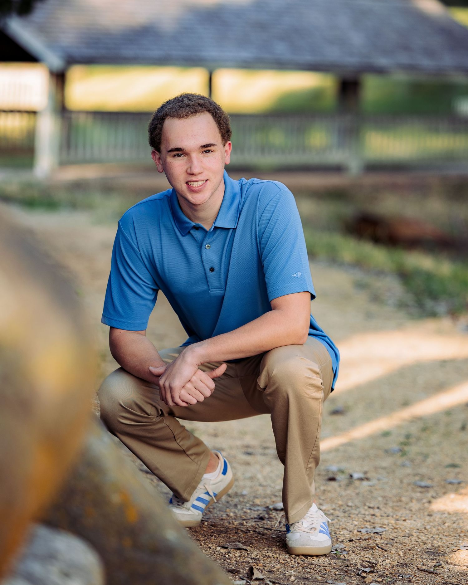 A young man in a blue polo shirt and khaki pants crouches outdoors for a senior portrait photo.