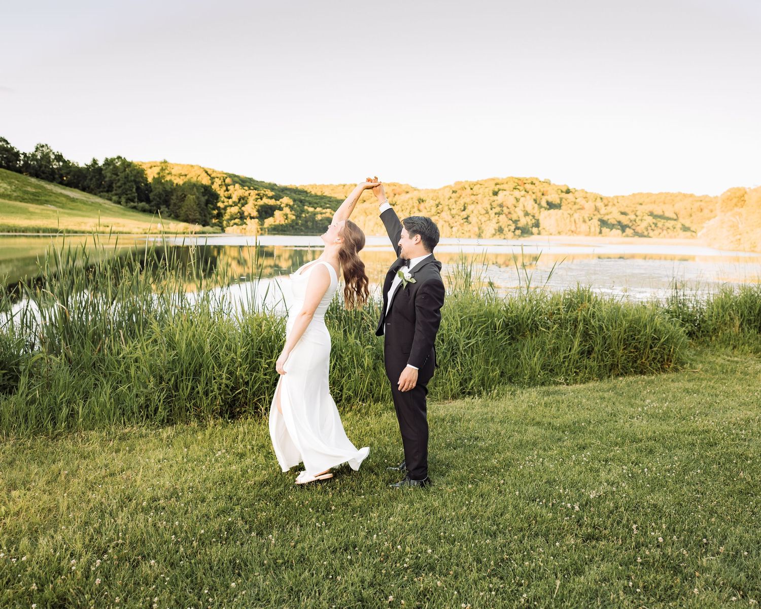 Bride and groom dancing by a scenic lake at golden hour, surrounded by lush greenery and rolling hills.