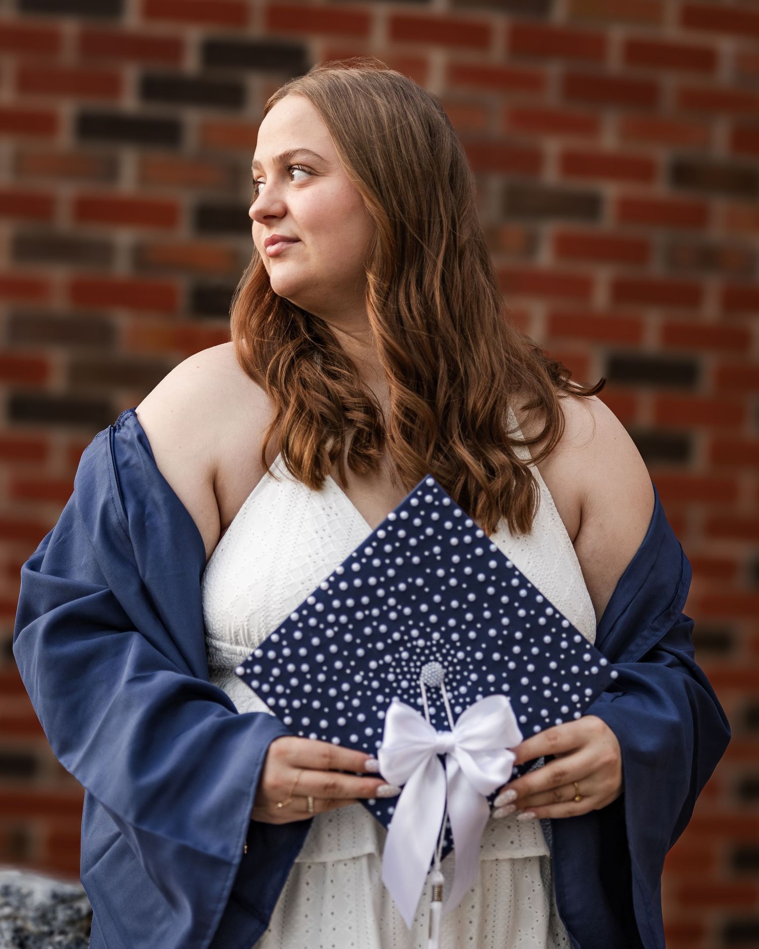 Young woman in white dress holding decorated navy graduation cap with white bow against brick wall.