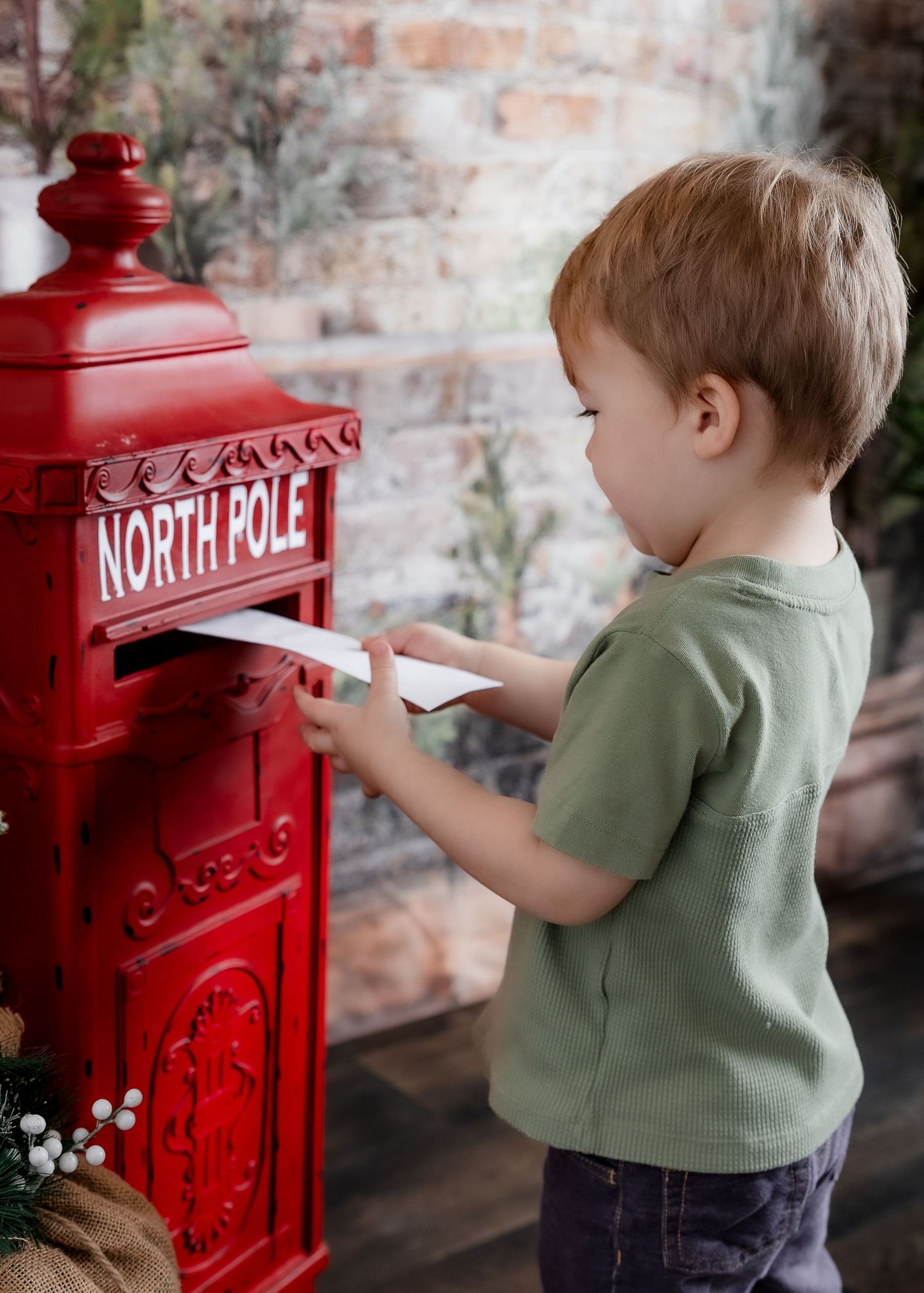 A young boy in a green shirt mailing a letter into a bright red North Pole mailbox during Christmas.