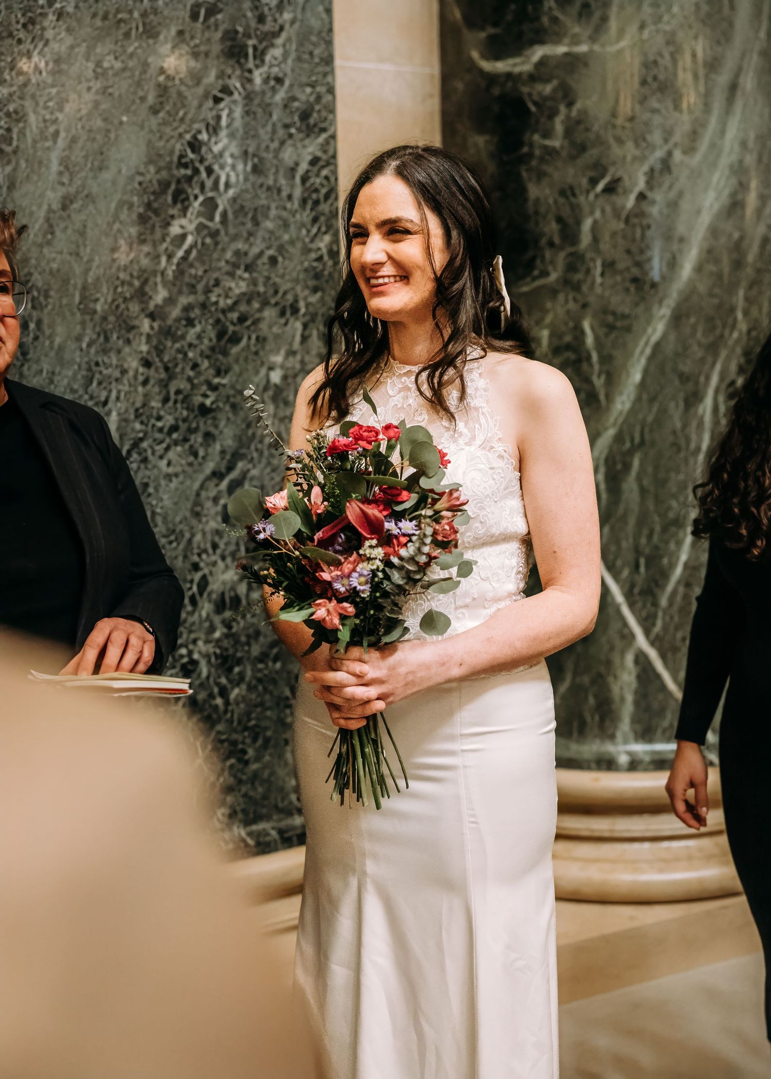Smiling bride in white lace gown holding vibrant red and pink bouquet during wedding ceremony.