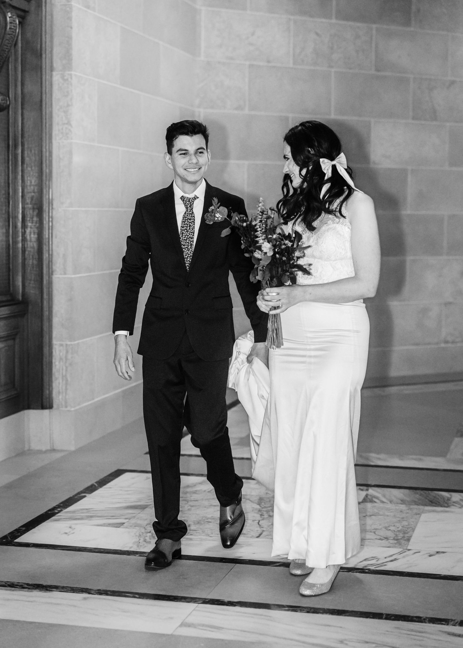 Smiling groom in black suit walks beside bride in white dress holding floral bouquet.