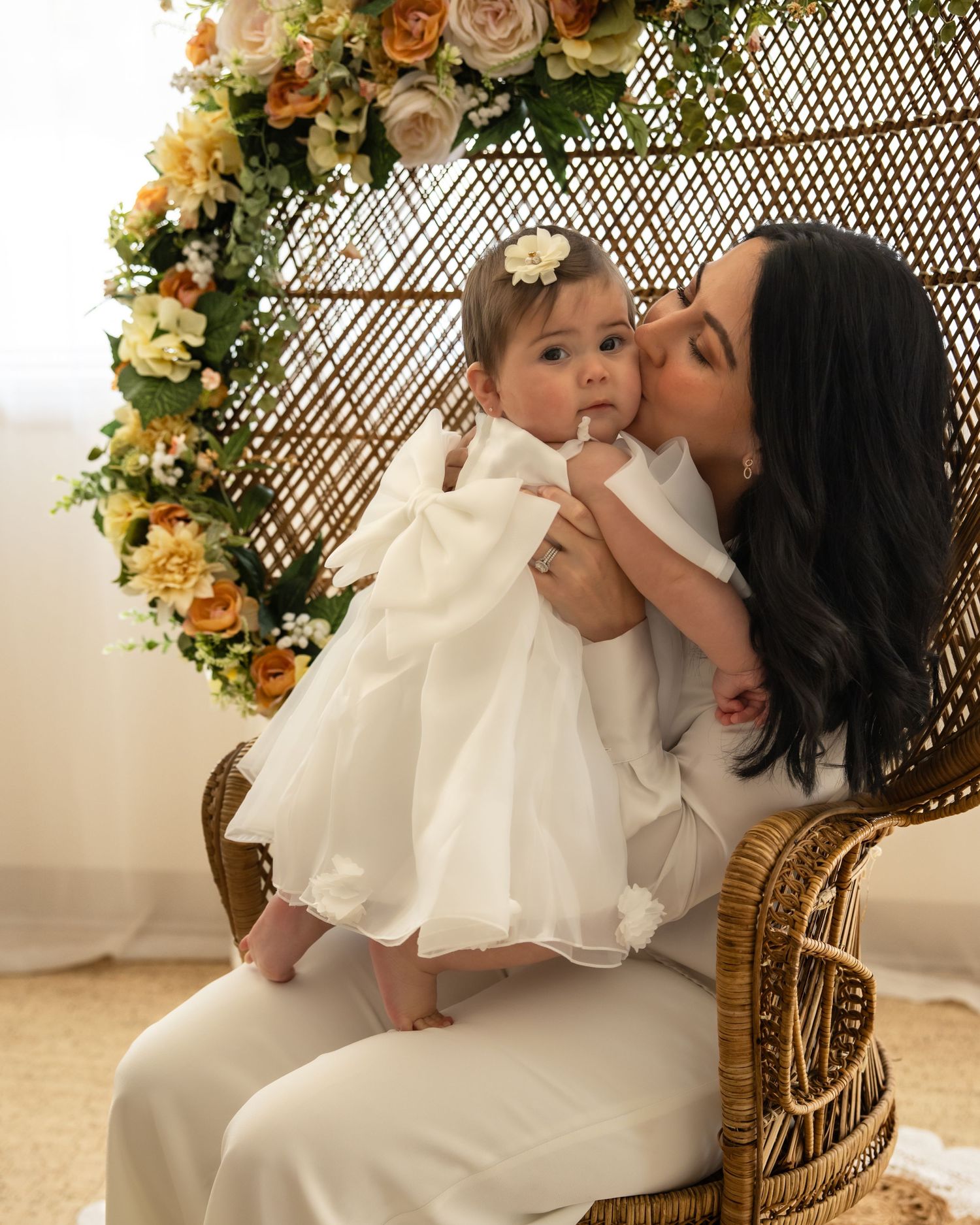 Dark-haired mother kissing baby girl in white dress while seated in a wicker peacock chair with flowers.