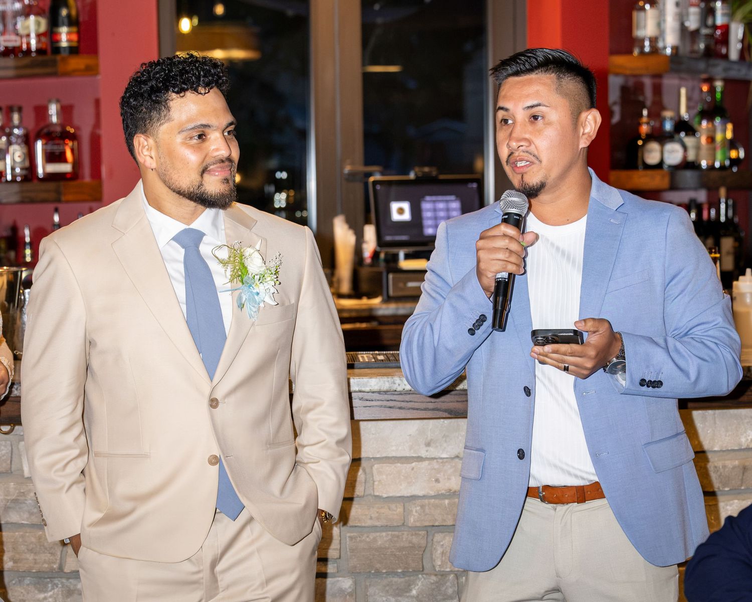 Groom in a tan suit with a boutonniere listens as a man in a blue blazer gives a microphone toast at a restaurant wedding reception.