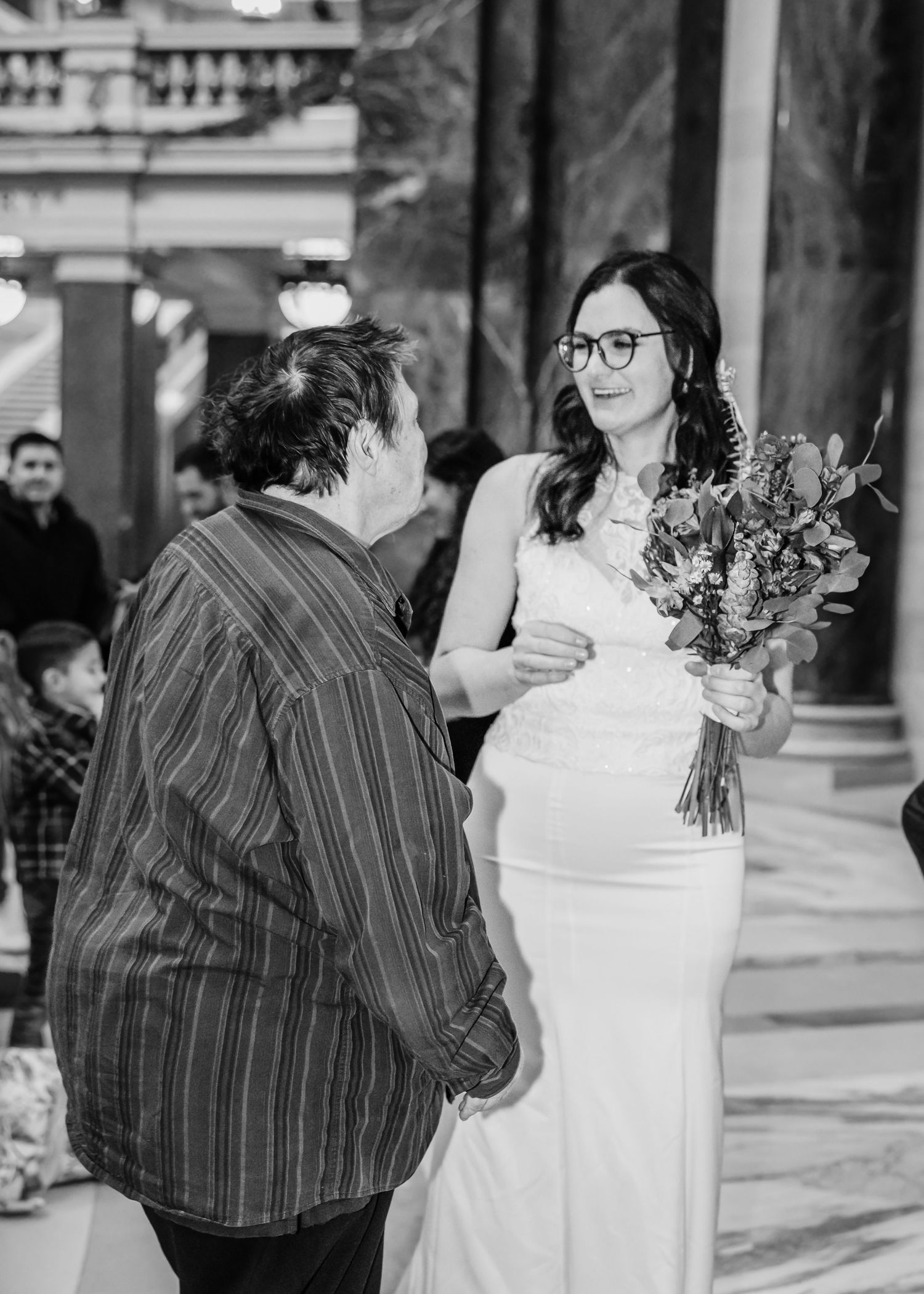 Bride in white gown holding a floral bouquet speaks with a guest at an elegant wedding venue.