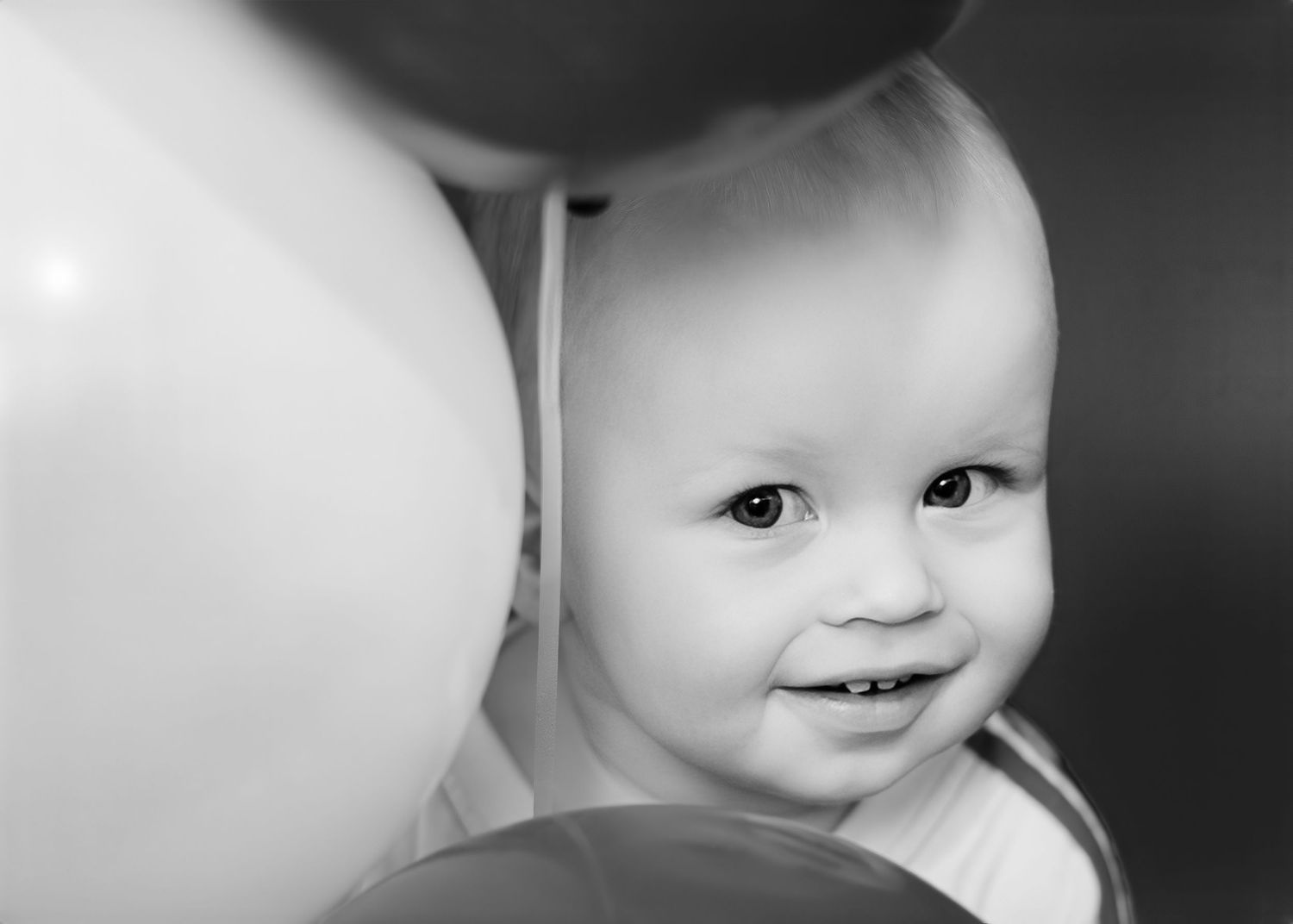 Black and white close-up portrait of smiling baby peeking through balloons.