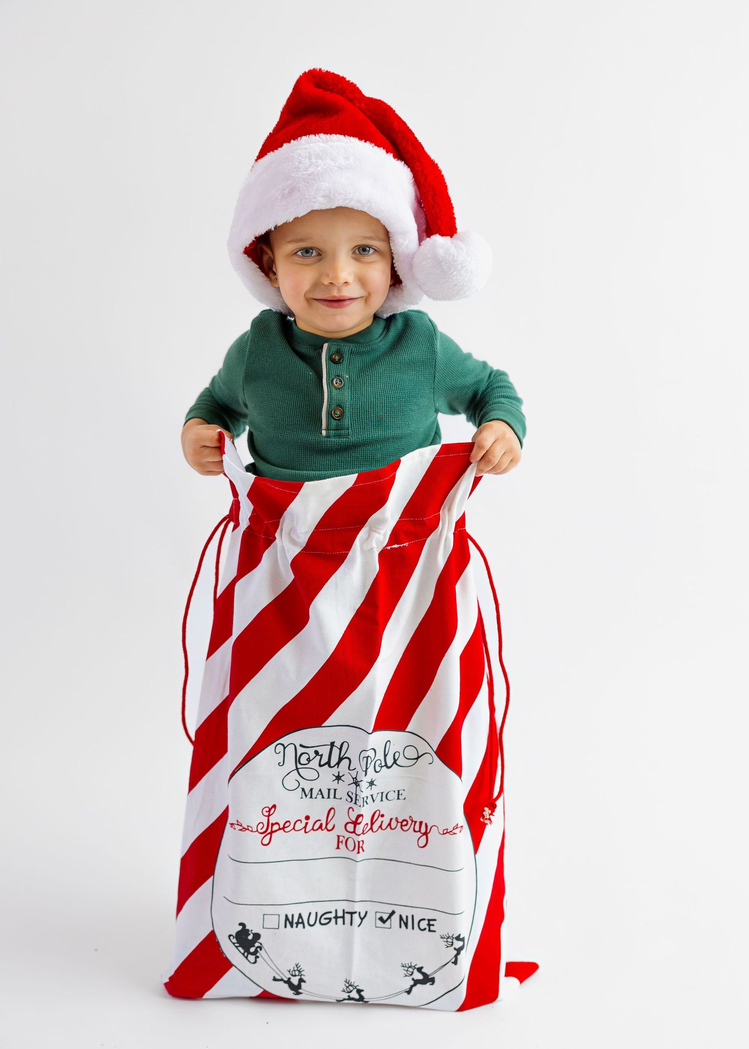 Happy toddler wearing Santa hat peeks out from a red and white striped North Pole gift sack.