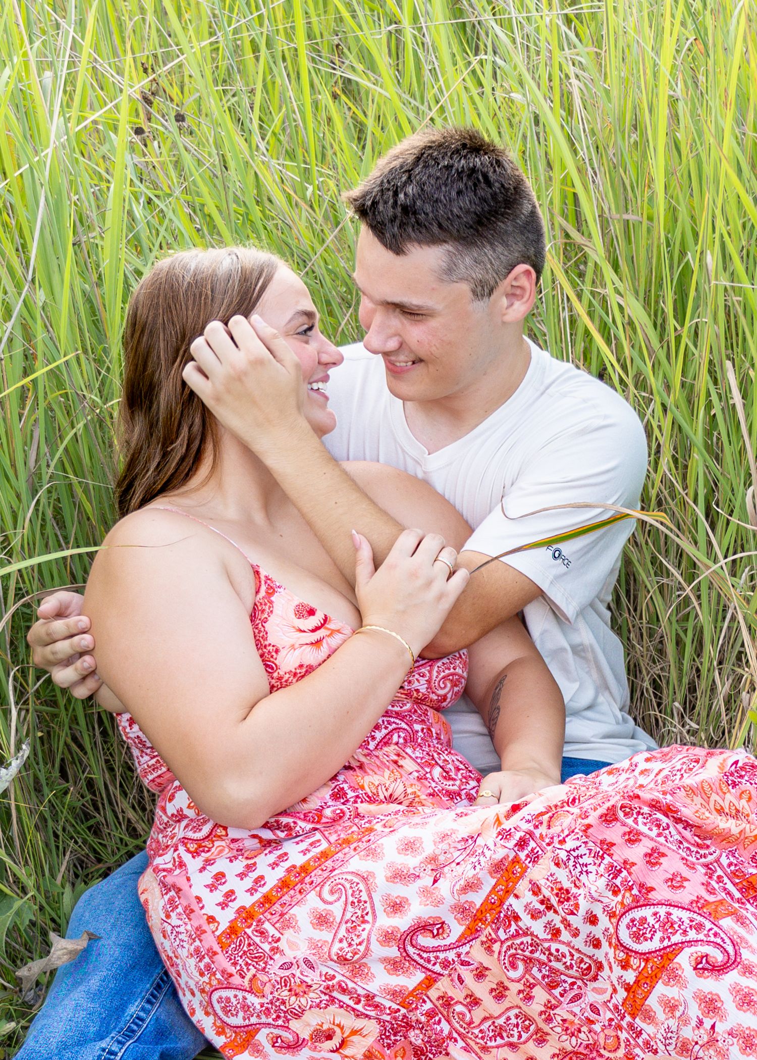 Romantic couple sitting in tall green grass, smiling at each other in a field.