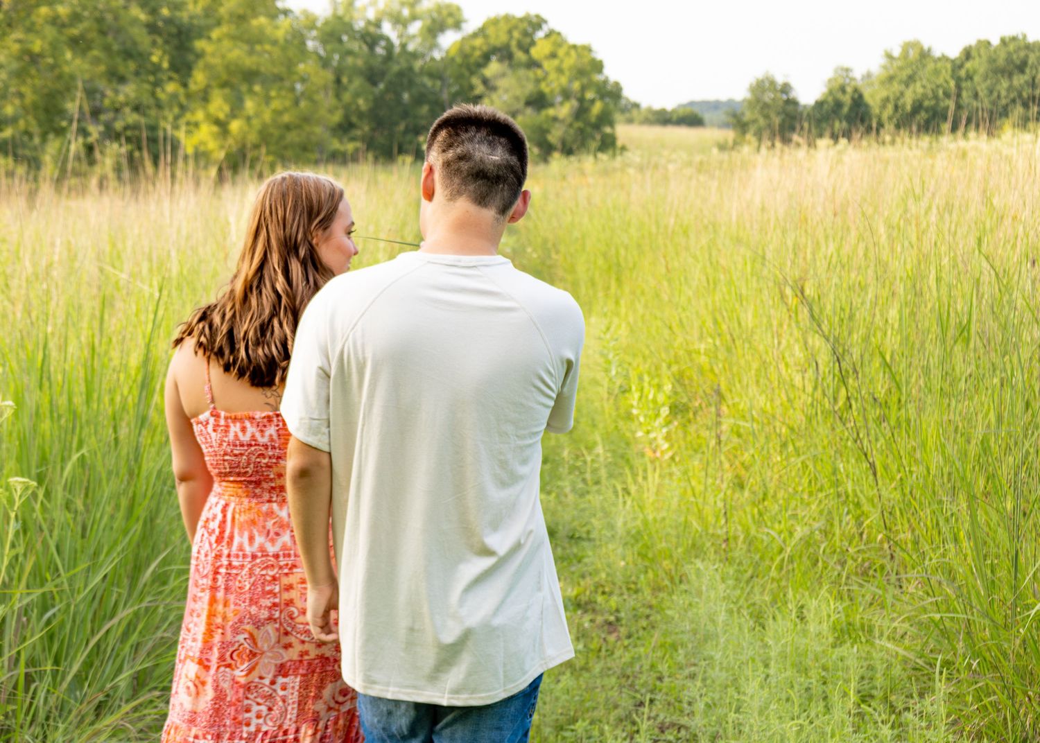 A couple stands hand-in-hand at the edge of a tall grass meadow, gazing into the field.