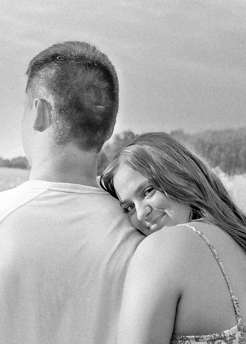 Black and white photo of a couple embracing outdoors, woman smiling over mans shoulder.