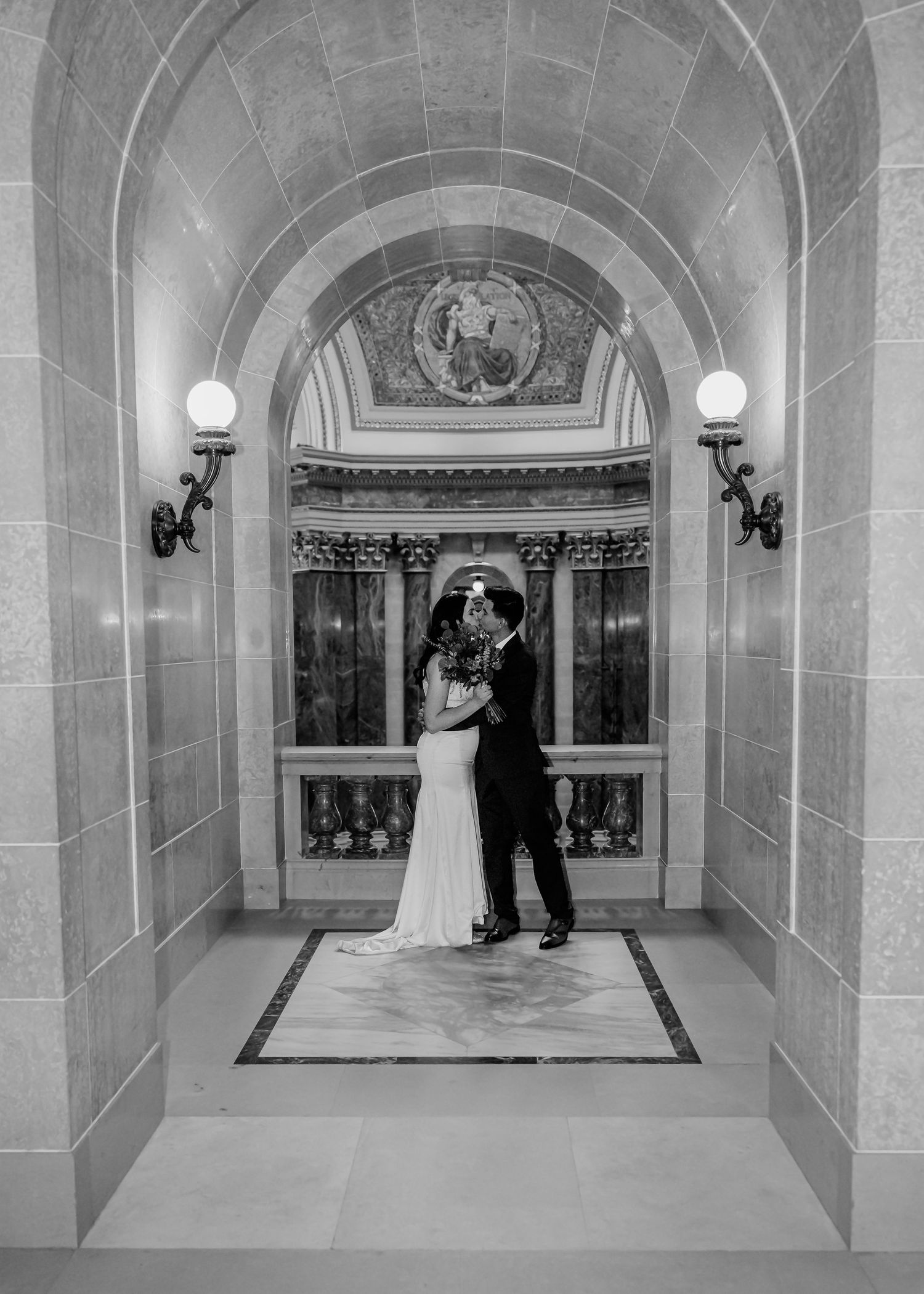Black and white photo of a bride and groom kissing under an ornate arched hallway.