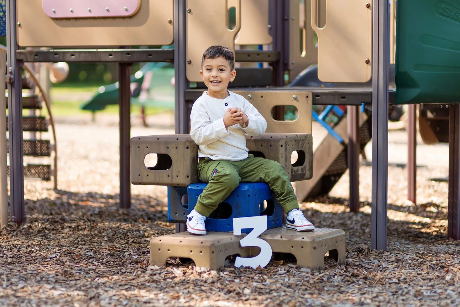 Young boy in white shirt and green pants sits on playground equipment holding number three sign.