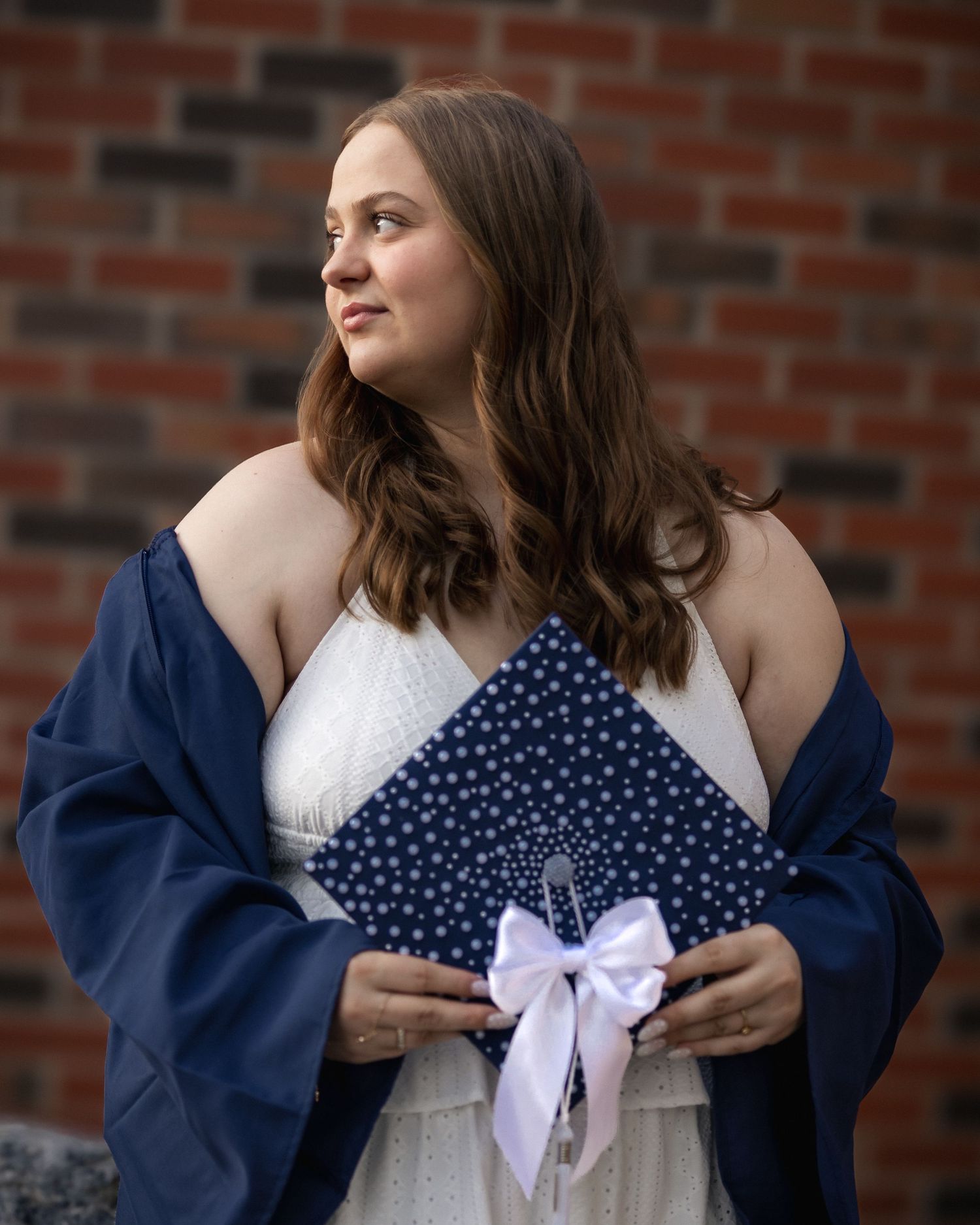Young woman in navy graduation gown holds decorated cap with white bow, posing outdoors near brick wall.